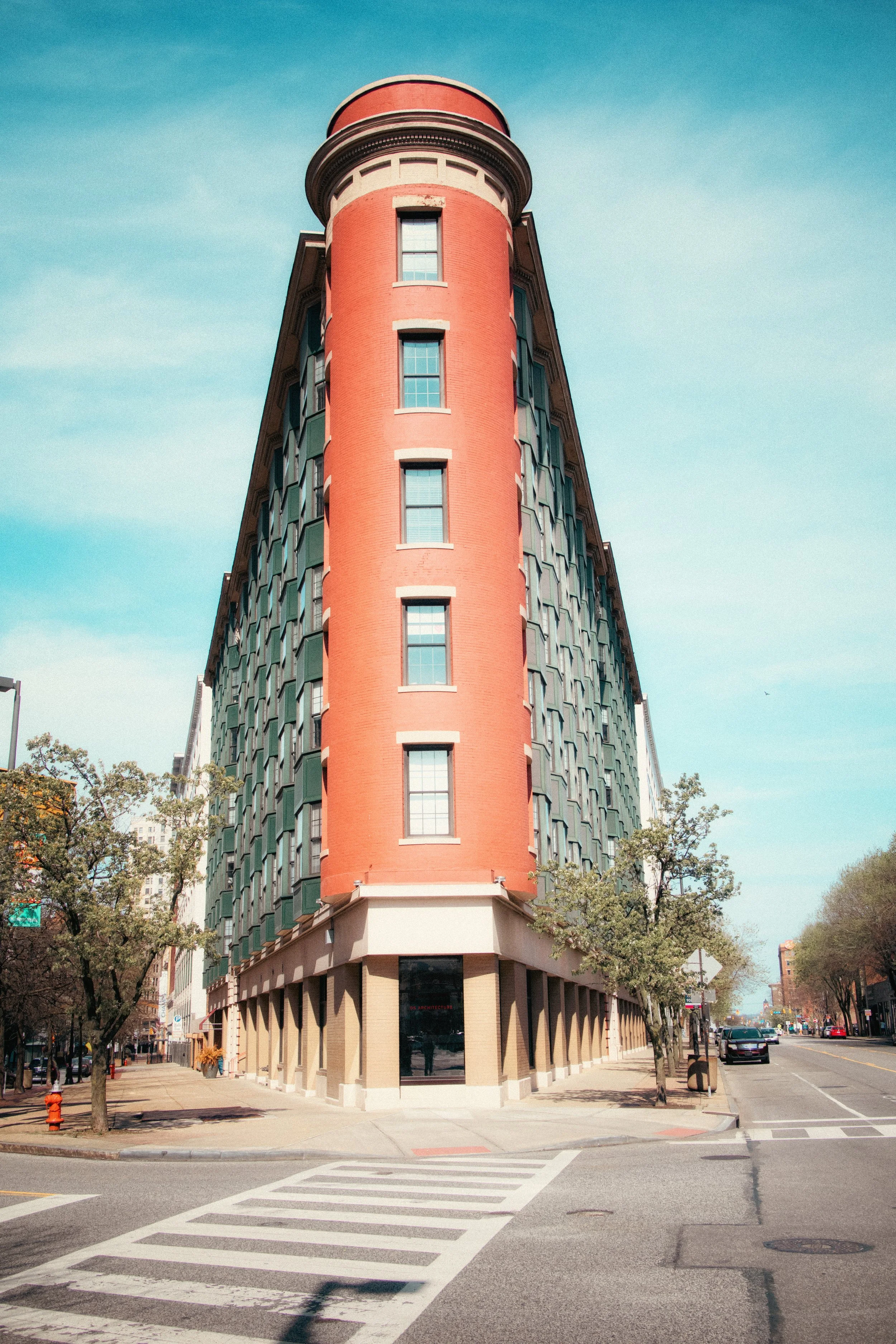 Corner of a red brick building with rounded turret, storefront at street level, in a city with trees and cars, clear daytime sky.