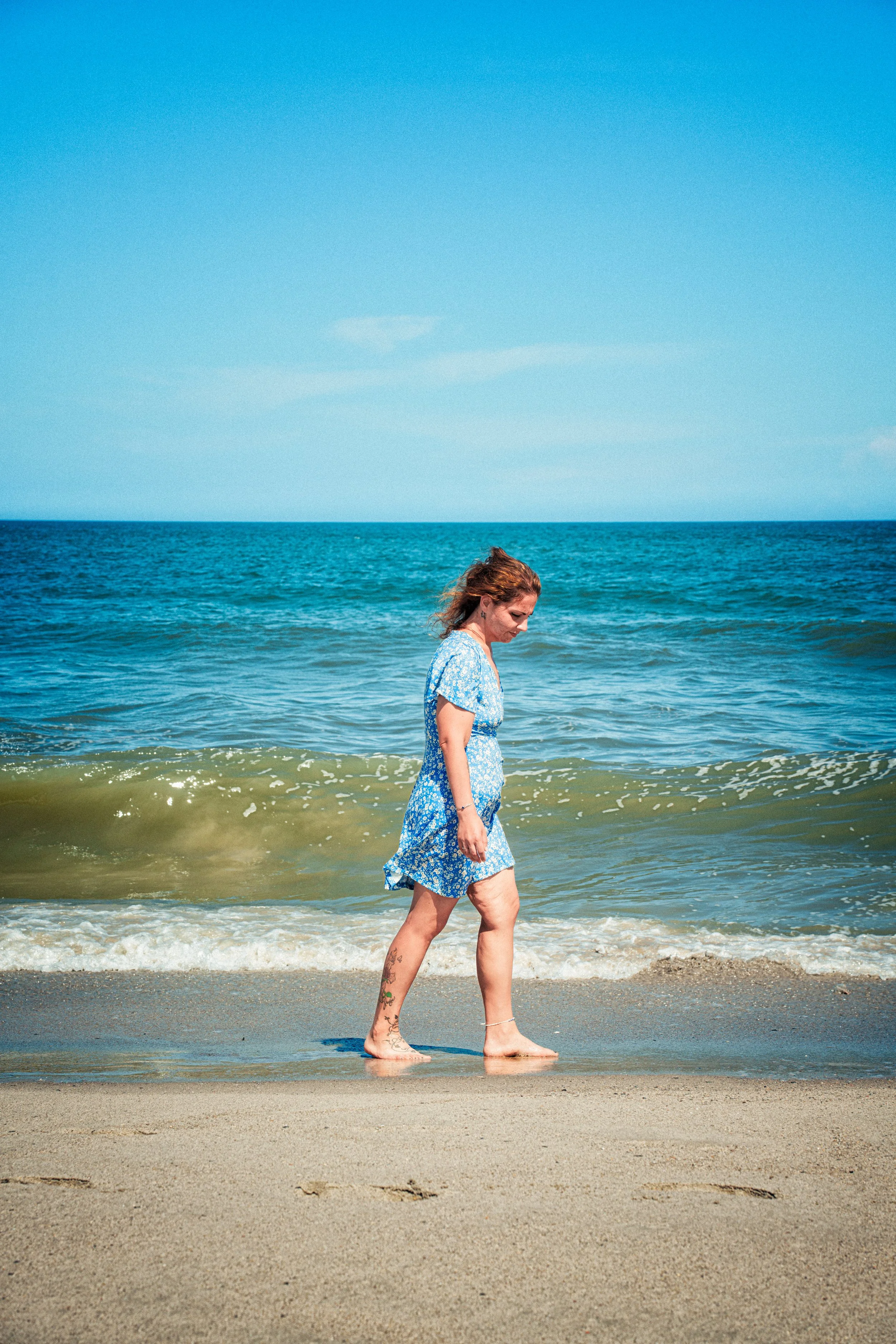 A woman walking along the shore of a beach with ocean waves in the background, wearing a blue floral dress and barefoot.