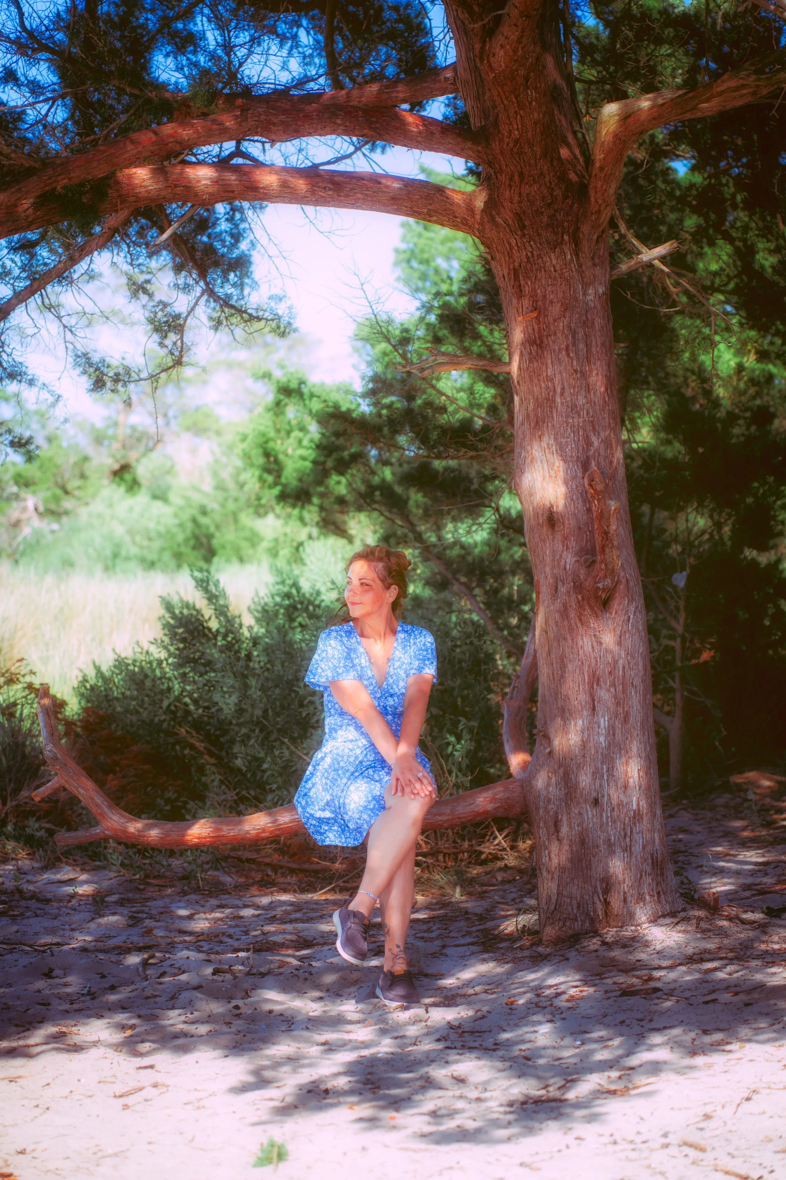 The subject stands among trees in warm natural light, capturing an unposed and reflective outdoor portrait.