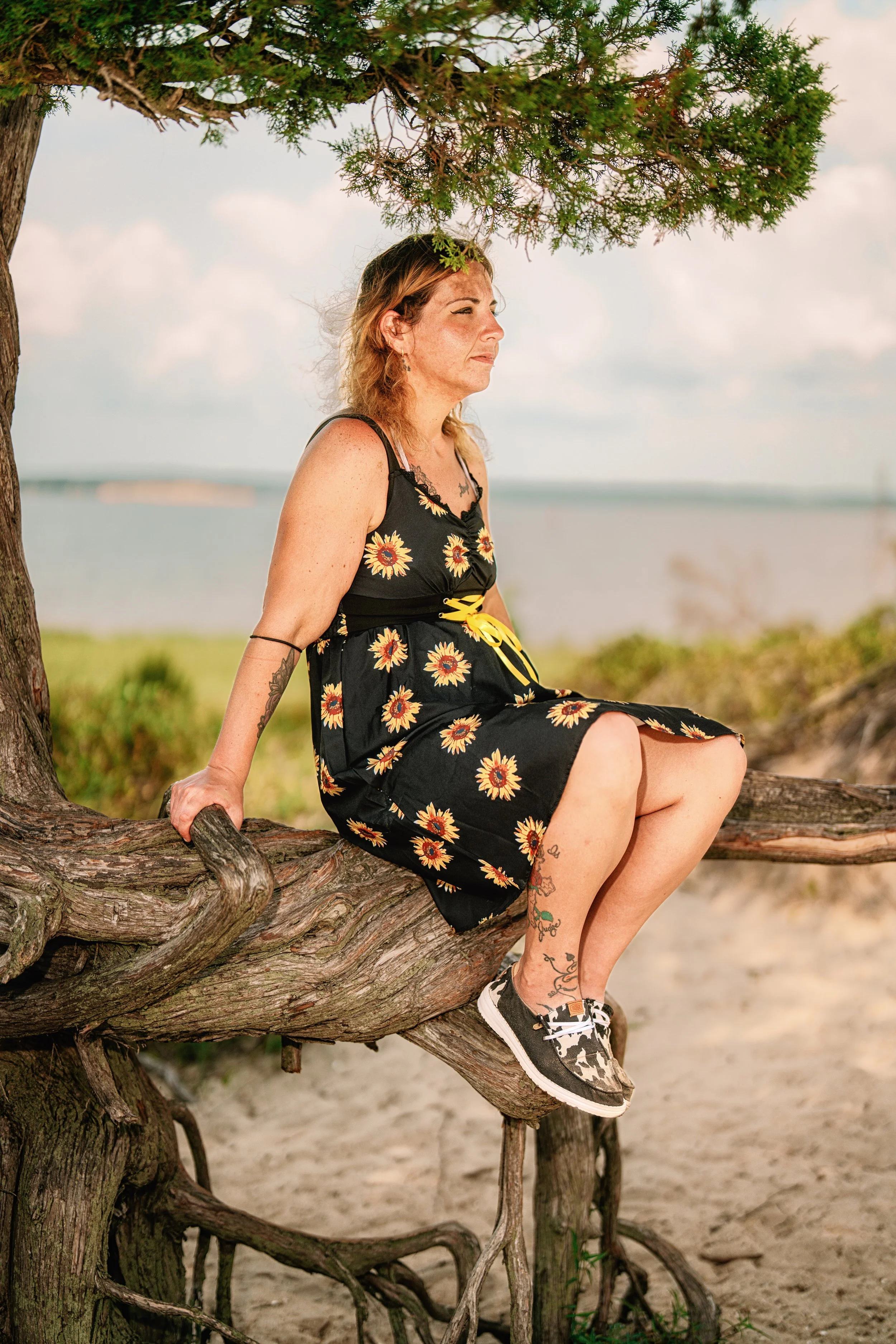 A woman with tattoos and curly, shoulder-length hair sitting on a large tree branch near the beach, under a green tree with a view of the water in the background.