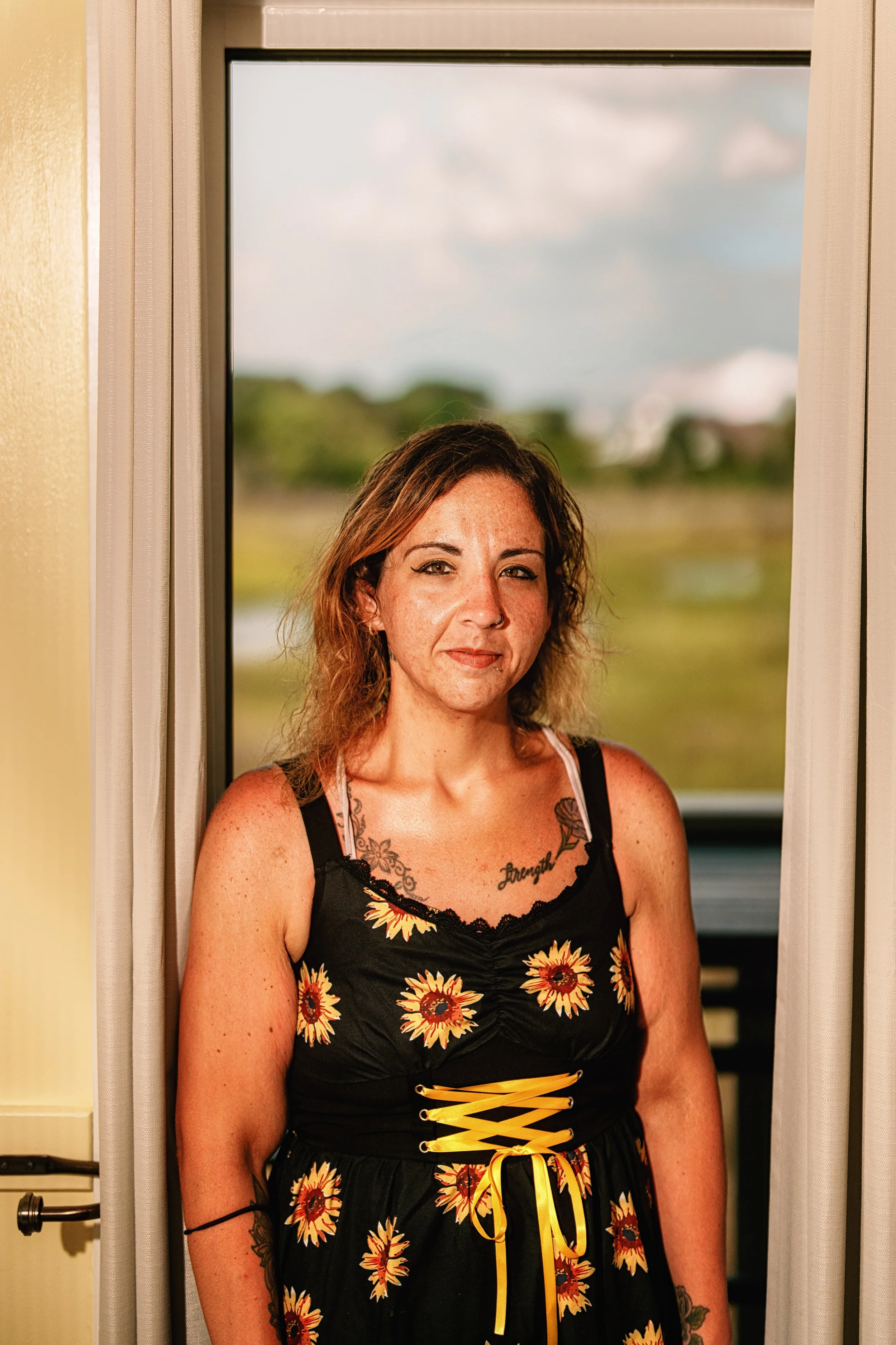A woman with shoulder-length brown curly hair and tattoos on her chest and arms, wearing a black floral dress with yellow laces, standing in front of a window with a landscape view.