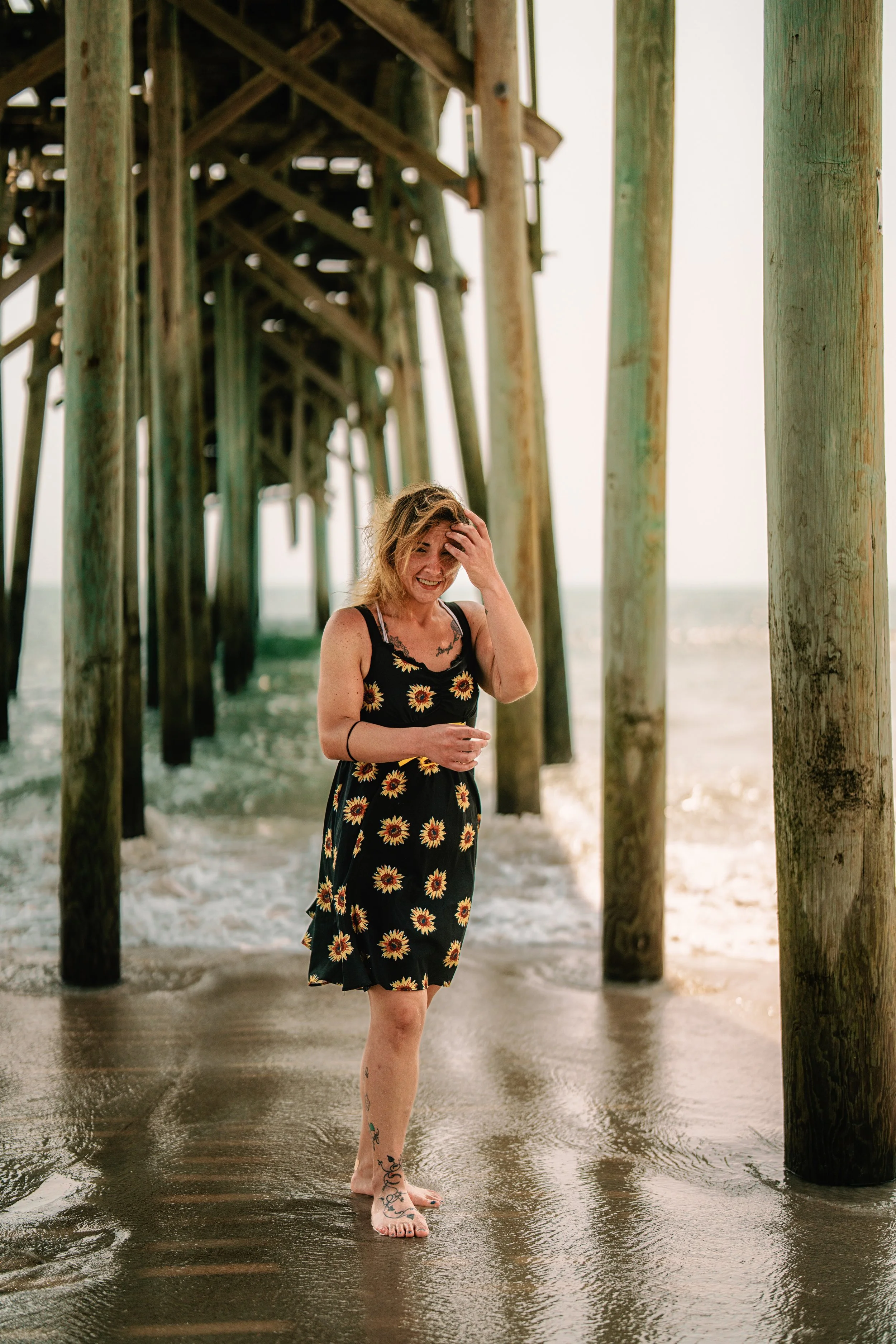 A woman standing barefoot on wet sand under a wooden pier, with her hand on her head and smiling. She is wearing a black dress with sunflower patterns and has tattoos on her legs. The background shows the ocean waves and the sky.
