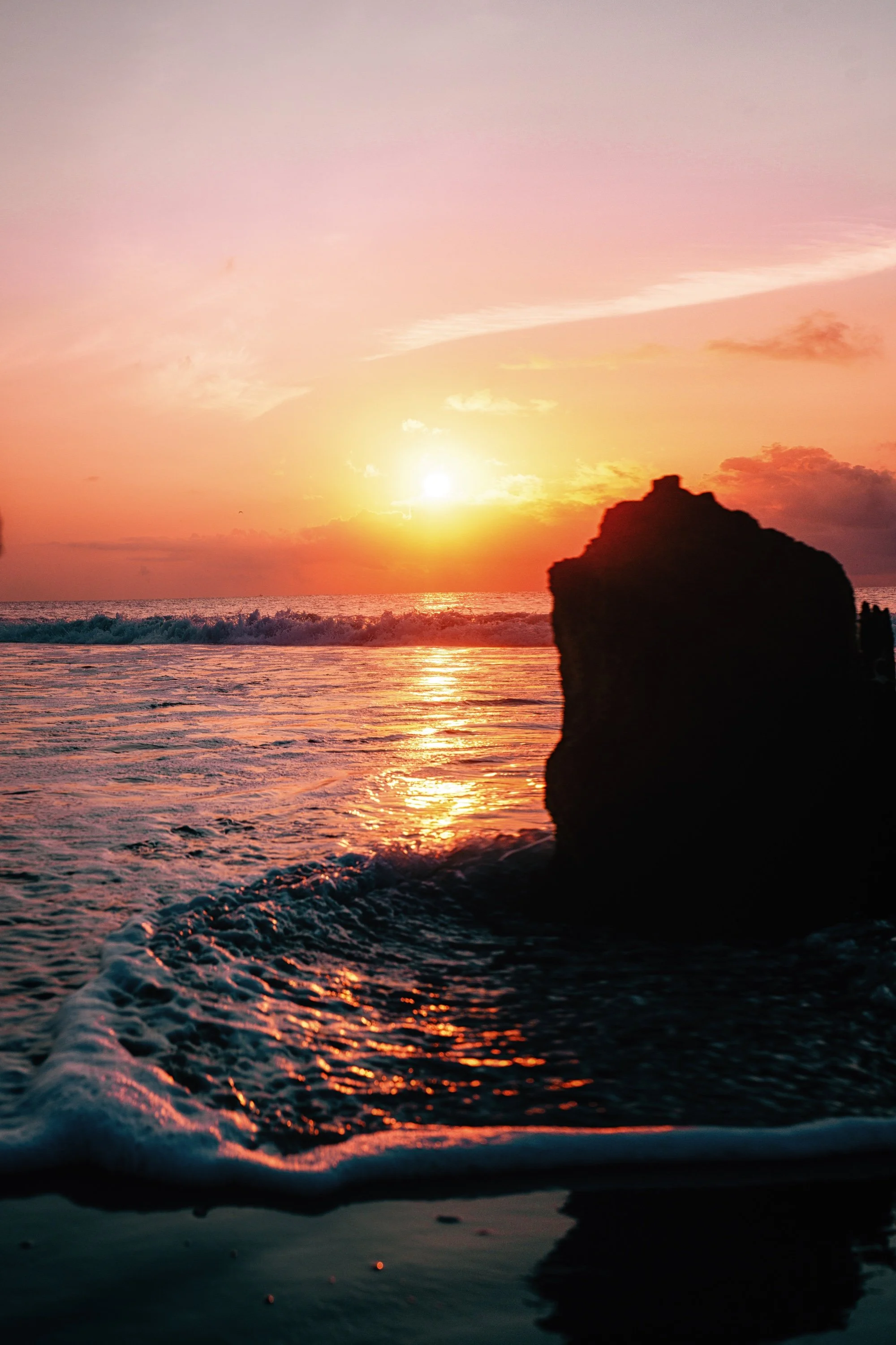 Sunset over the ocean with colorful pink, orange, and purple sky and a silhouette of a rock formation in the foreground.