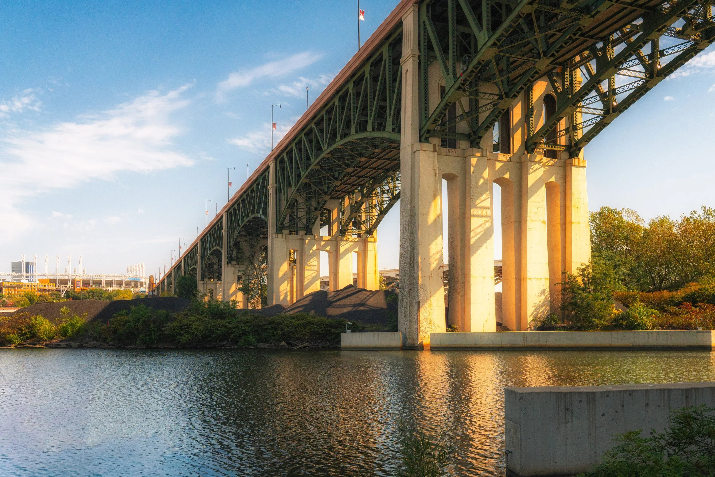 A large bridge with green metal framework crossing over a body of water, with a cityscape in the background and trees on the shoreline, during sunset.