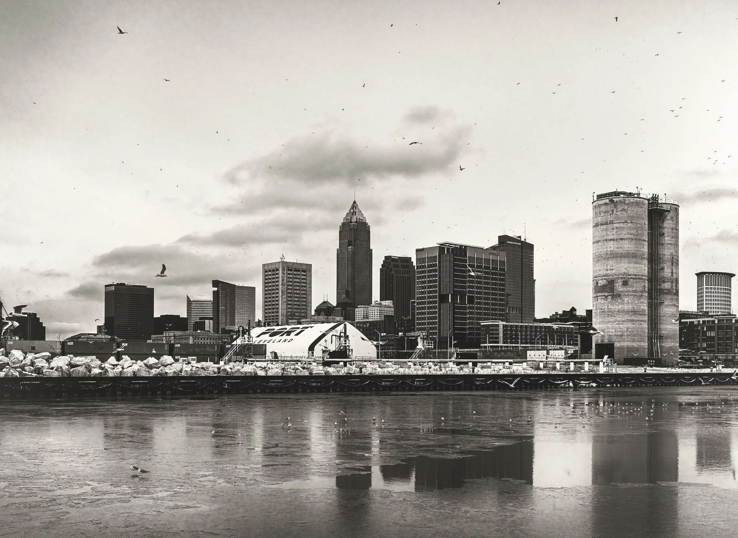 The Cleveland skyline stretches across calm water, rendered in black and white to emphasize form, contrast, and atmosphere.