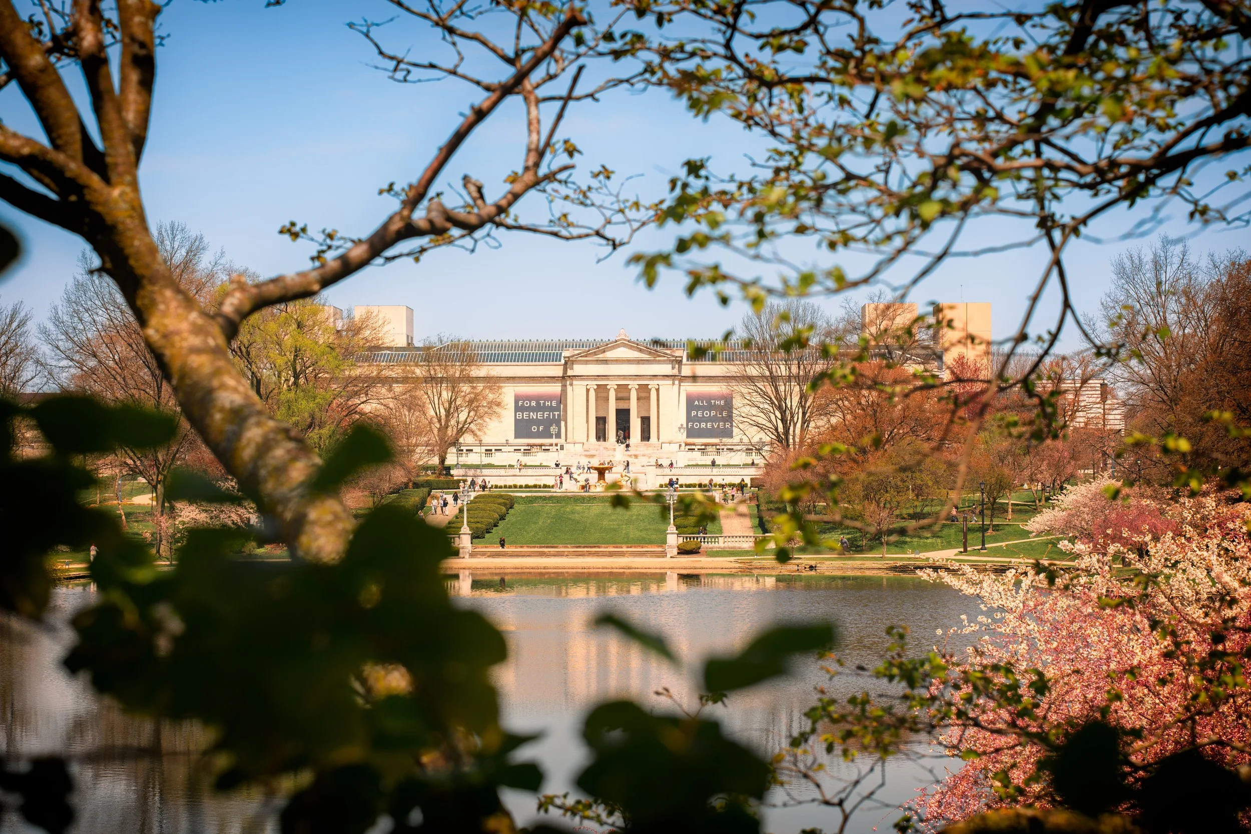 The image shows a view of a large classical building with columns, surrounded by trees and pink flowering trees, with a body of water in the foreground, during spring. The building has banners that read 'For the benefit of all the people forever'.