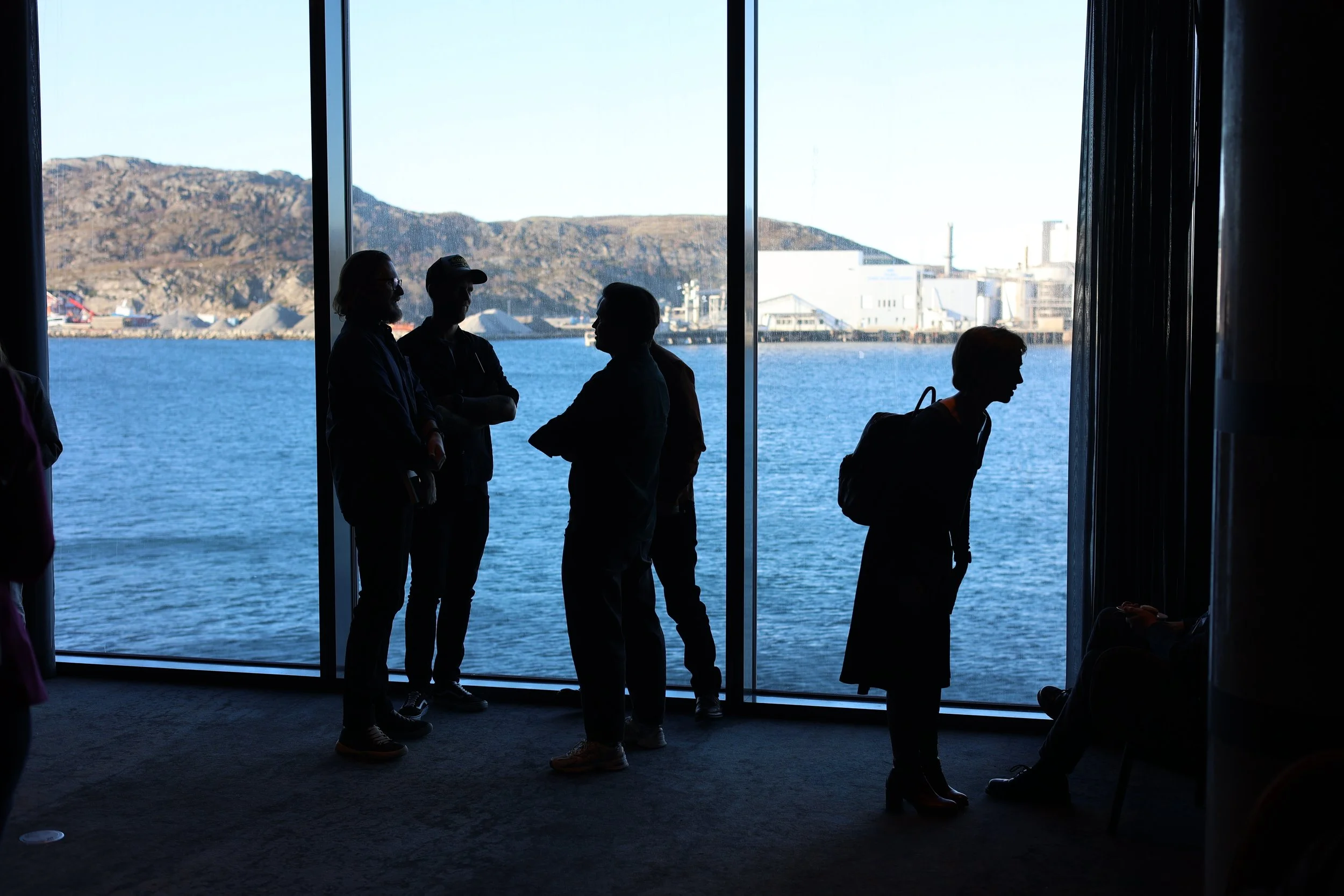 Silhuetter av mennesker som står ved et vindu med utsikt over vann og fjell i bakgrunnen.