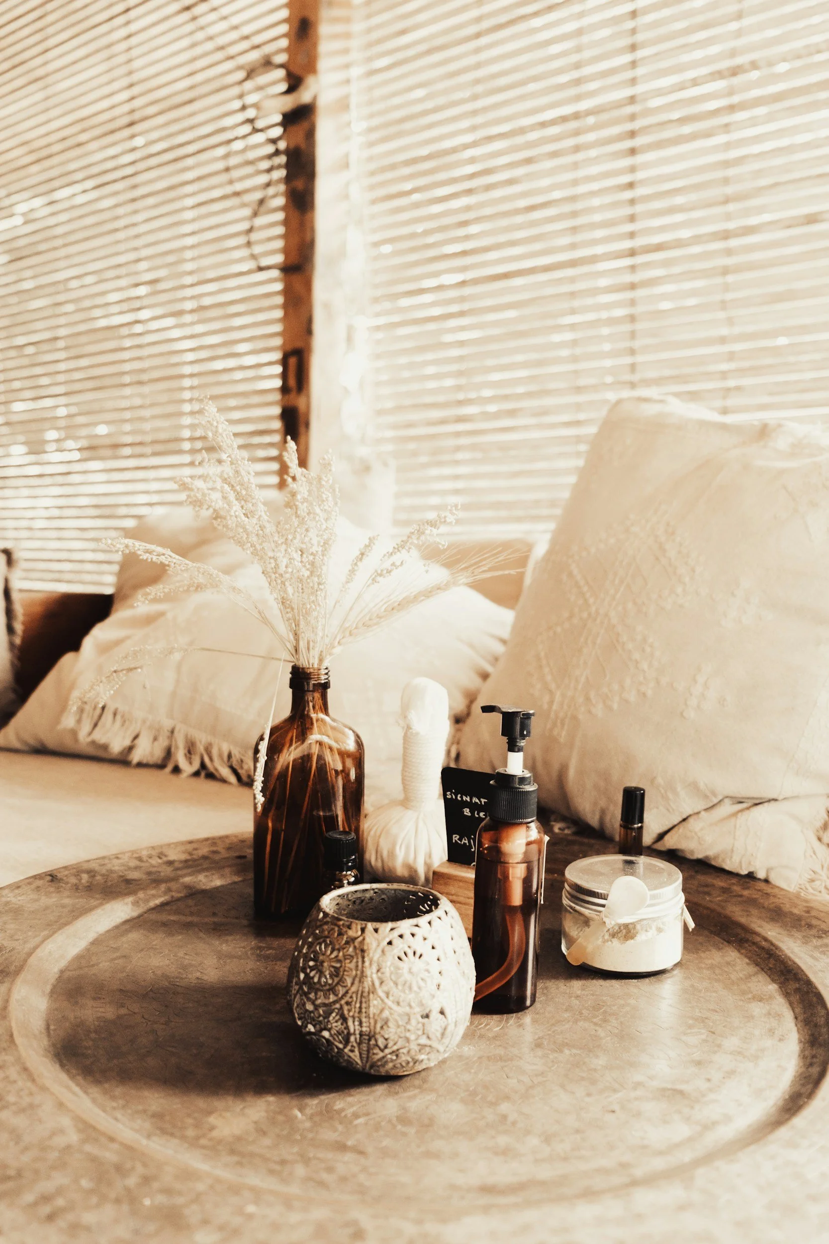 Decorative tray on a bed with a brown vase holding dried white flowers, a black pump bottle, a small jar, a textured white spherical object, and a small blackboard with writing. In the background, there are white pillows and closed wooden blinds with sunlight filtering through.