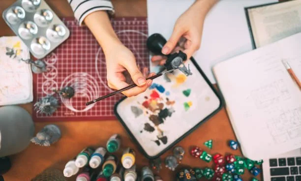 Person holding a paintbrush and a figurine while working with paints, dice, stationery, and a laptop on a desk.