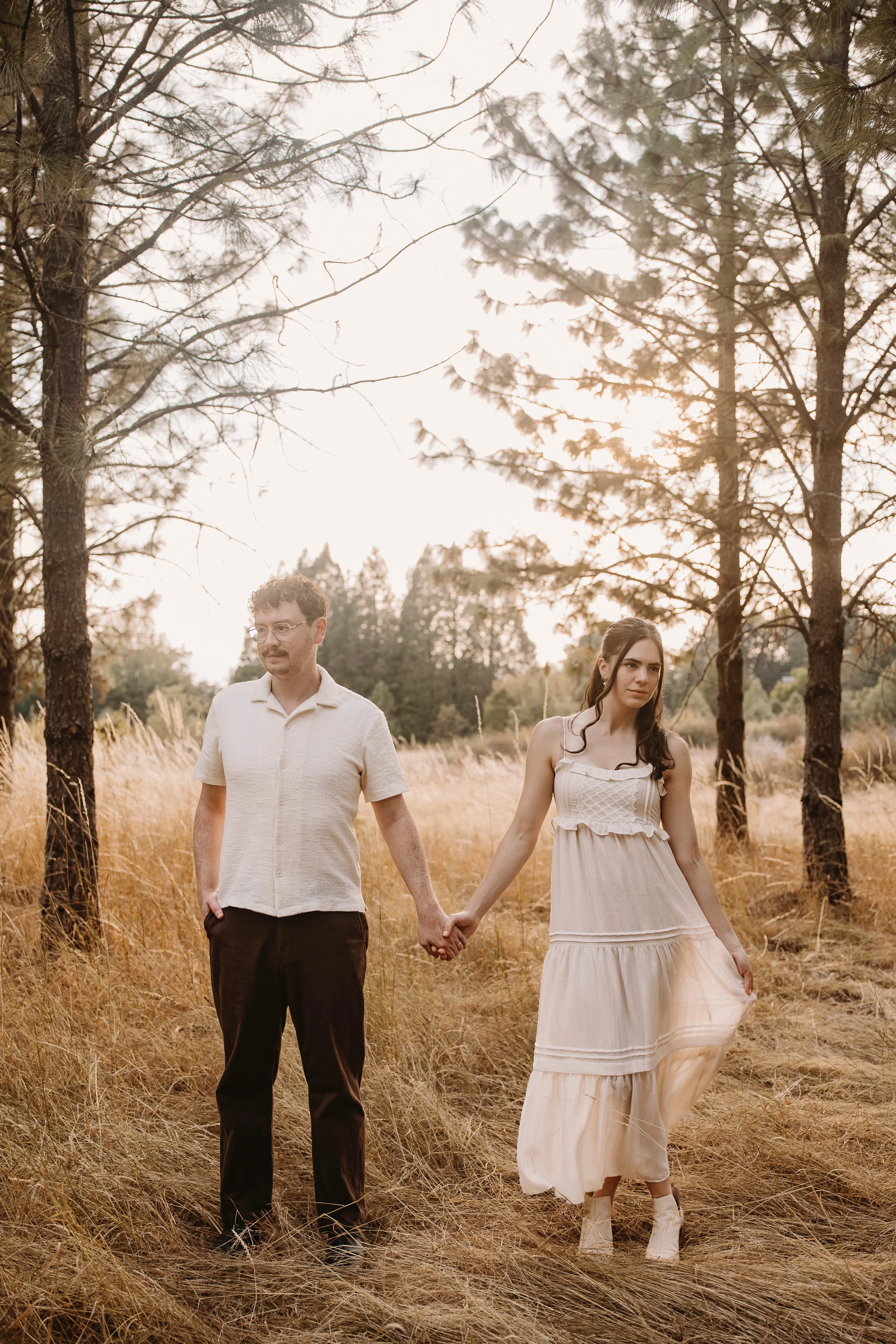 A young couple holding hands walking through a grassy field with trees in the background, sunlight shining through the trees during sunset or sunrise.