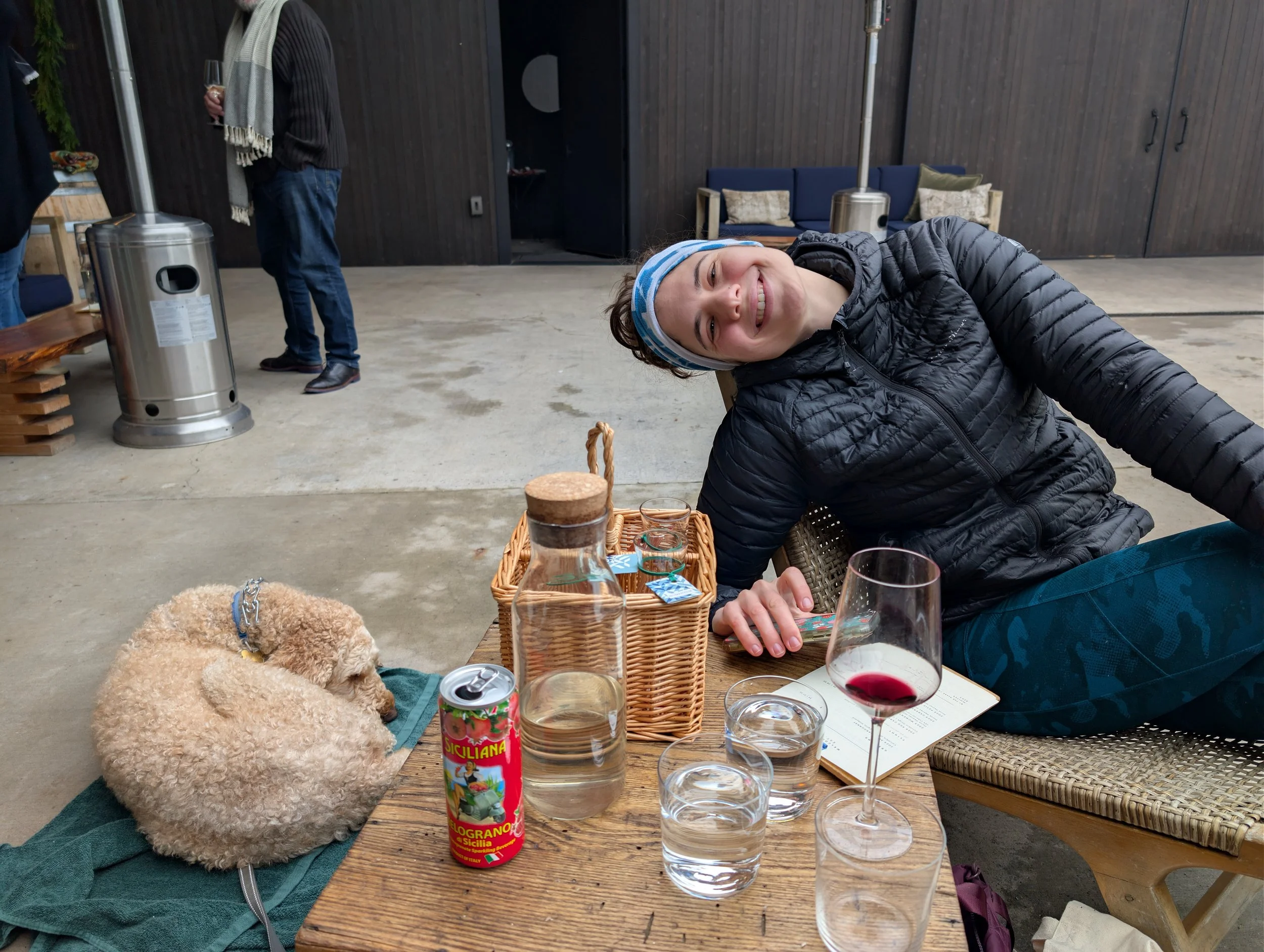A young woman in a black jacket and blue headband smiling while reclining at a table with a dog, wine glass, and beverages at an outdoor gathering.