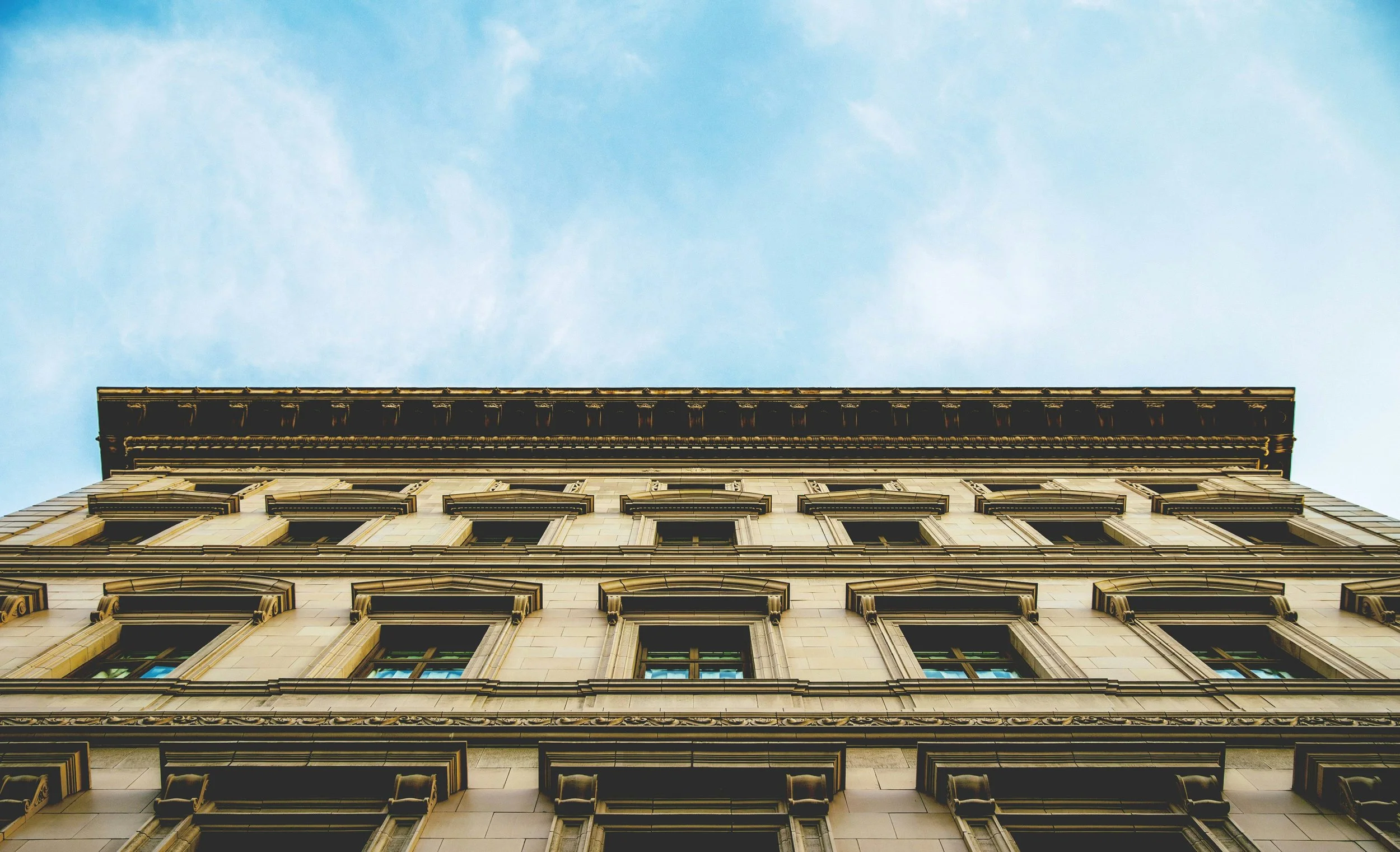 Upward view of a classical building with multiple windows and decorative architectural details, against a blue sky with wispy clouds.