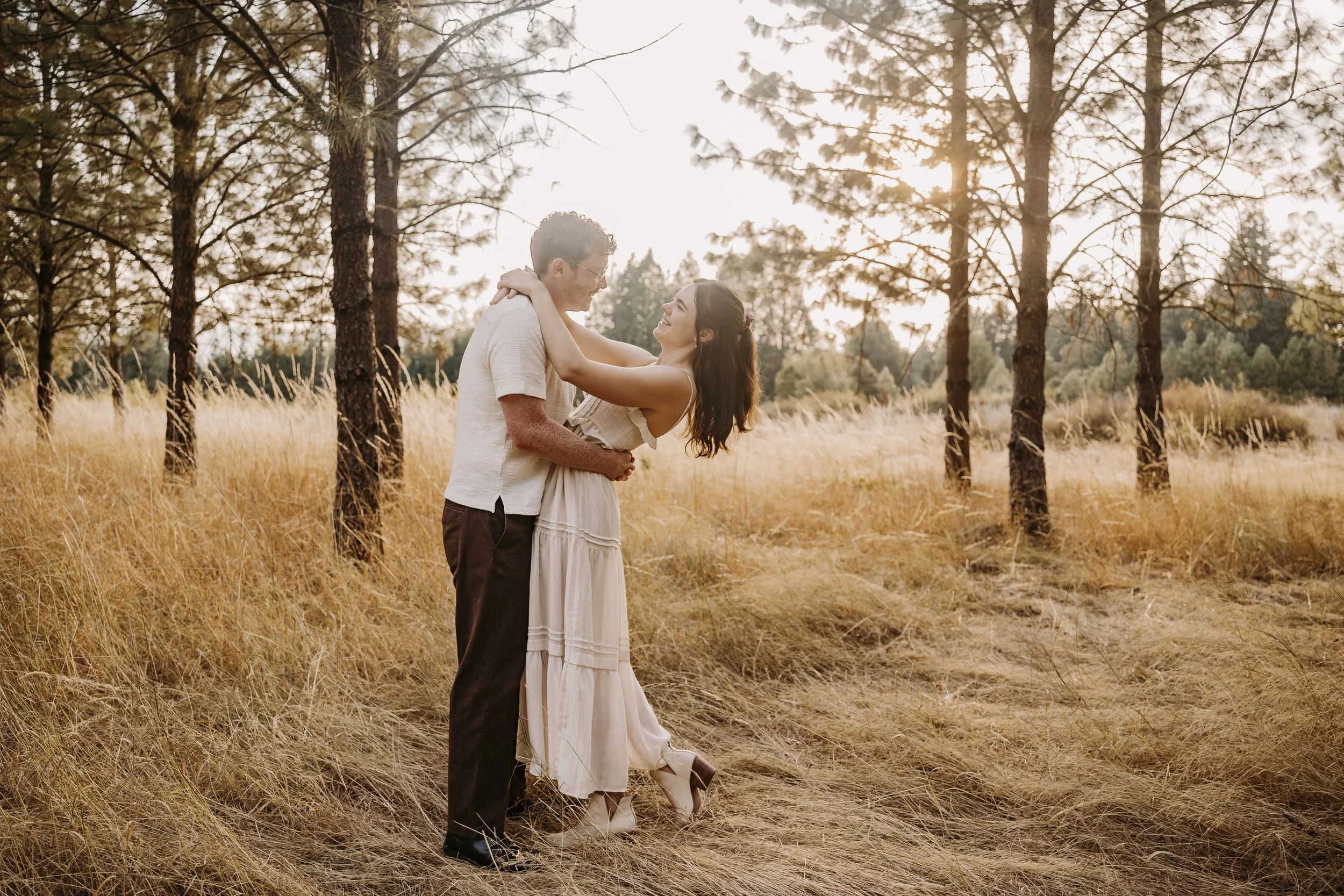 A couple standing in a forest clearing during sunset, with the man holding the woman as she leans towards him, smiling, with the golden light filtering through the trees.