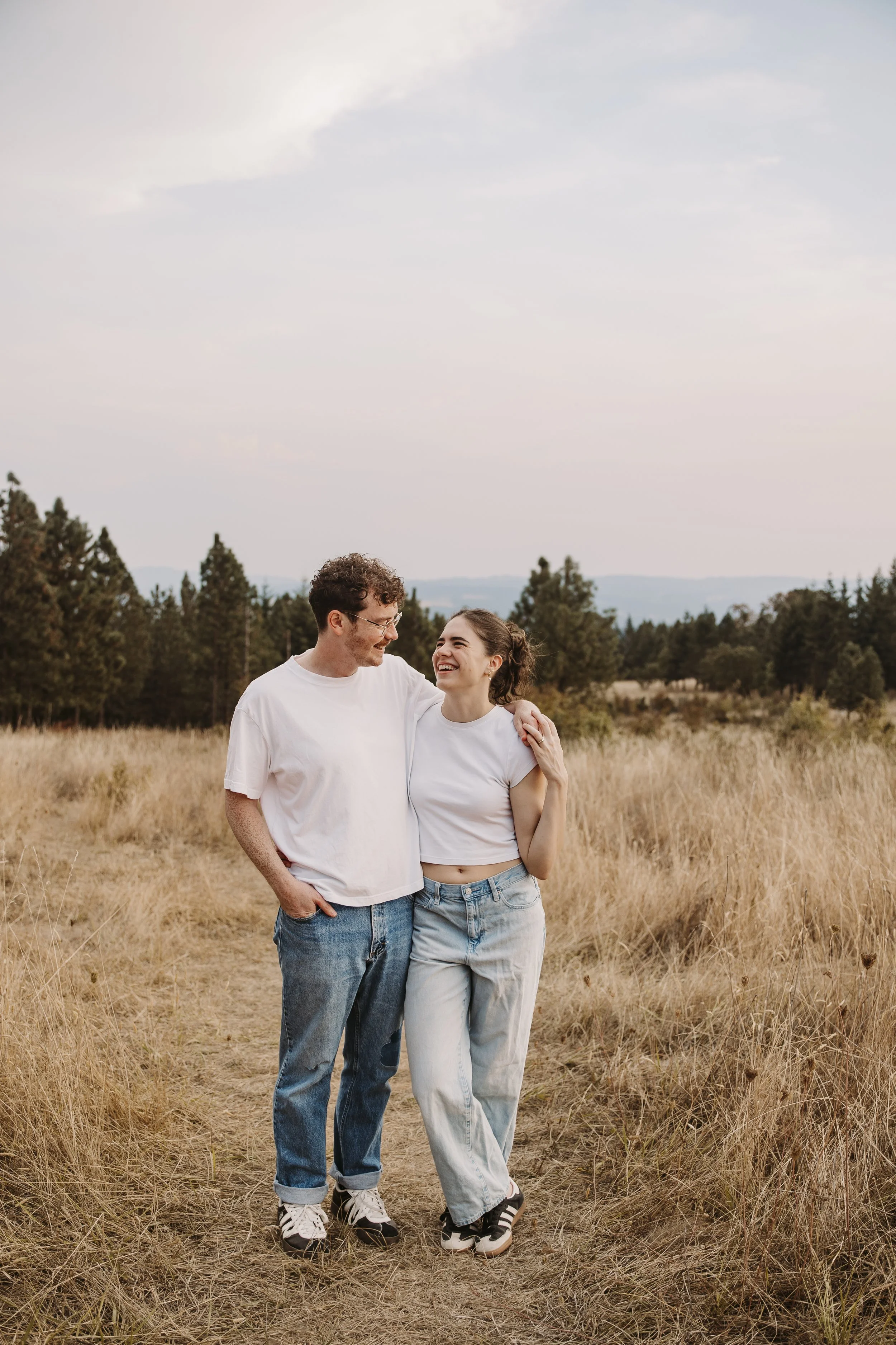 A young couple walking together in a field of tall, golden grass, smiling and enjoying each other's company on a clear day with a partly cloudy sky.