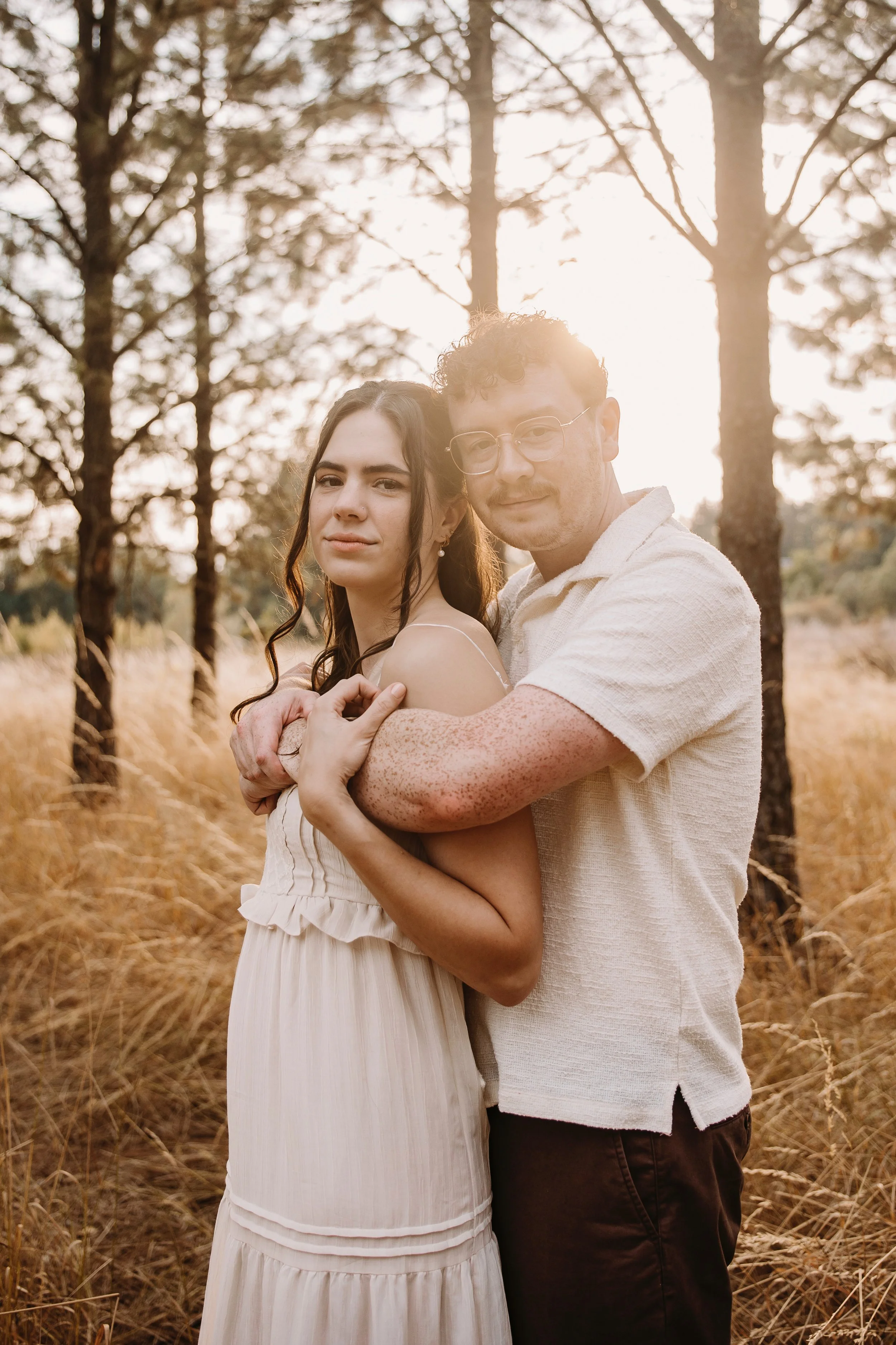 A young couple embracing in a forest with tall trees and golden grass, backlit by the setting sun.