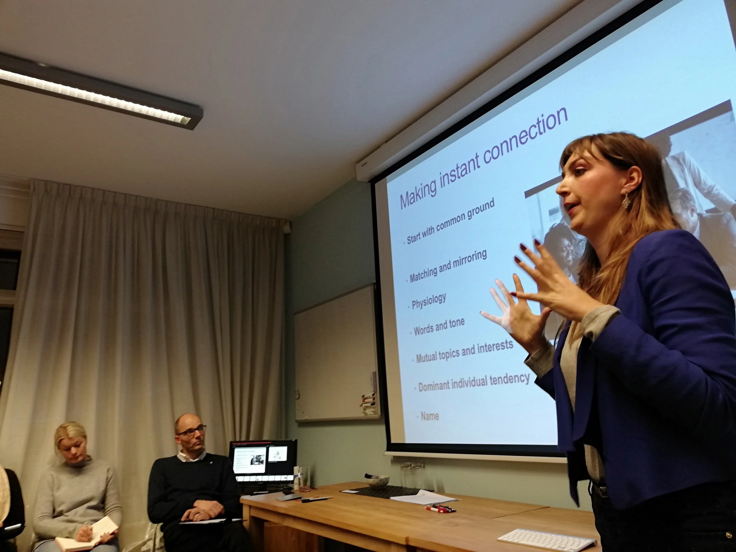 A woman giving a presentation in a conference room, with a slide titled 'Making instant connection' projected behind her. Three people are sitting at a table listening, while other participants are standing.