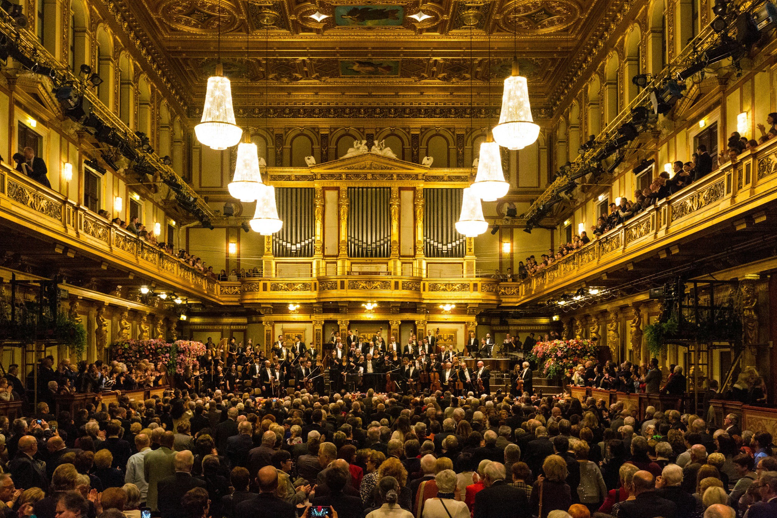 An elegant concert hall filled with audience members, with a choir and orchestra performing on stage. The hall is decorated with gold accents, chandeliers, and ornate architectural details.