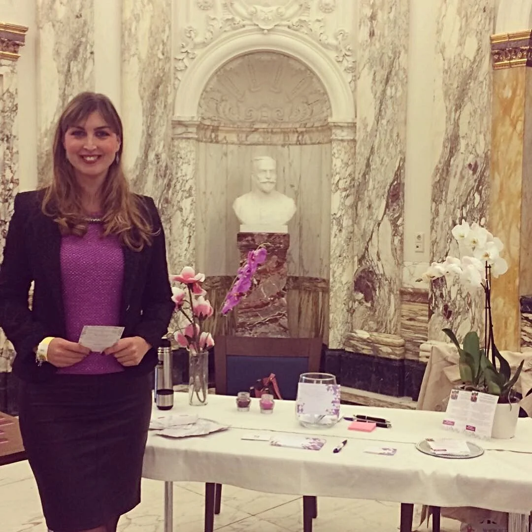 A woman smiling and holding papers stands next to a table with pink carnations, purple orchids, and various items, in a marble-lined room with ornate decor and a bust sculpture in the background.