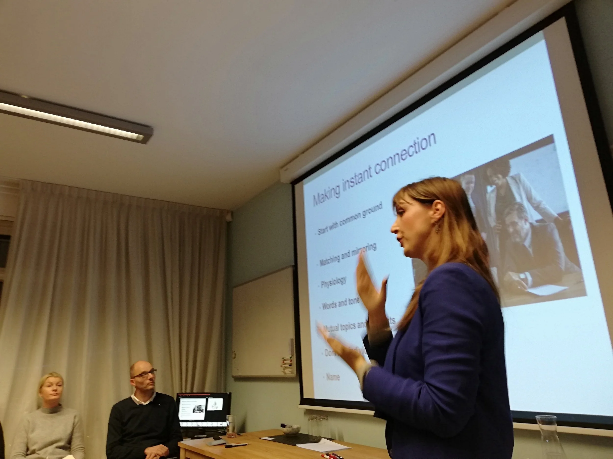 A woman in a purple blazer is giving a presentation in a conference room while standing next to a large projection screen. The screen displays a slide titled "Making instant connection" with bullet points. Several adults are seated at a table, listen