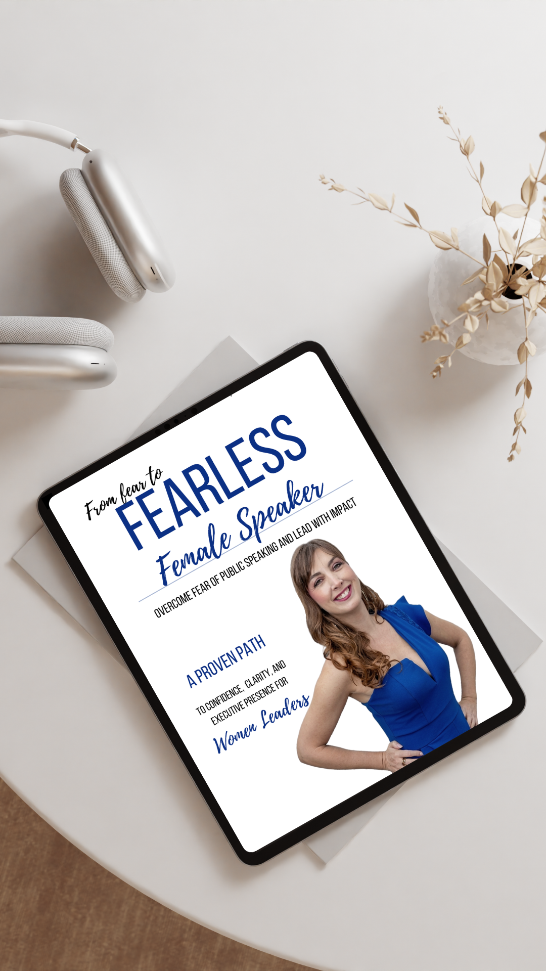 Tablet displaying a book titled "From fear to FEARLESS Female Speaker" on a white table next to a pair of white headphones and a white vase with dried beige leaves.