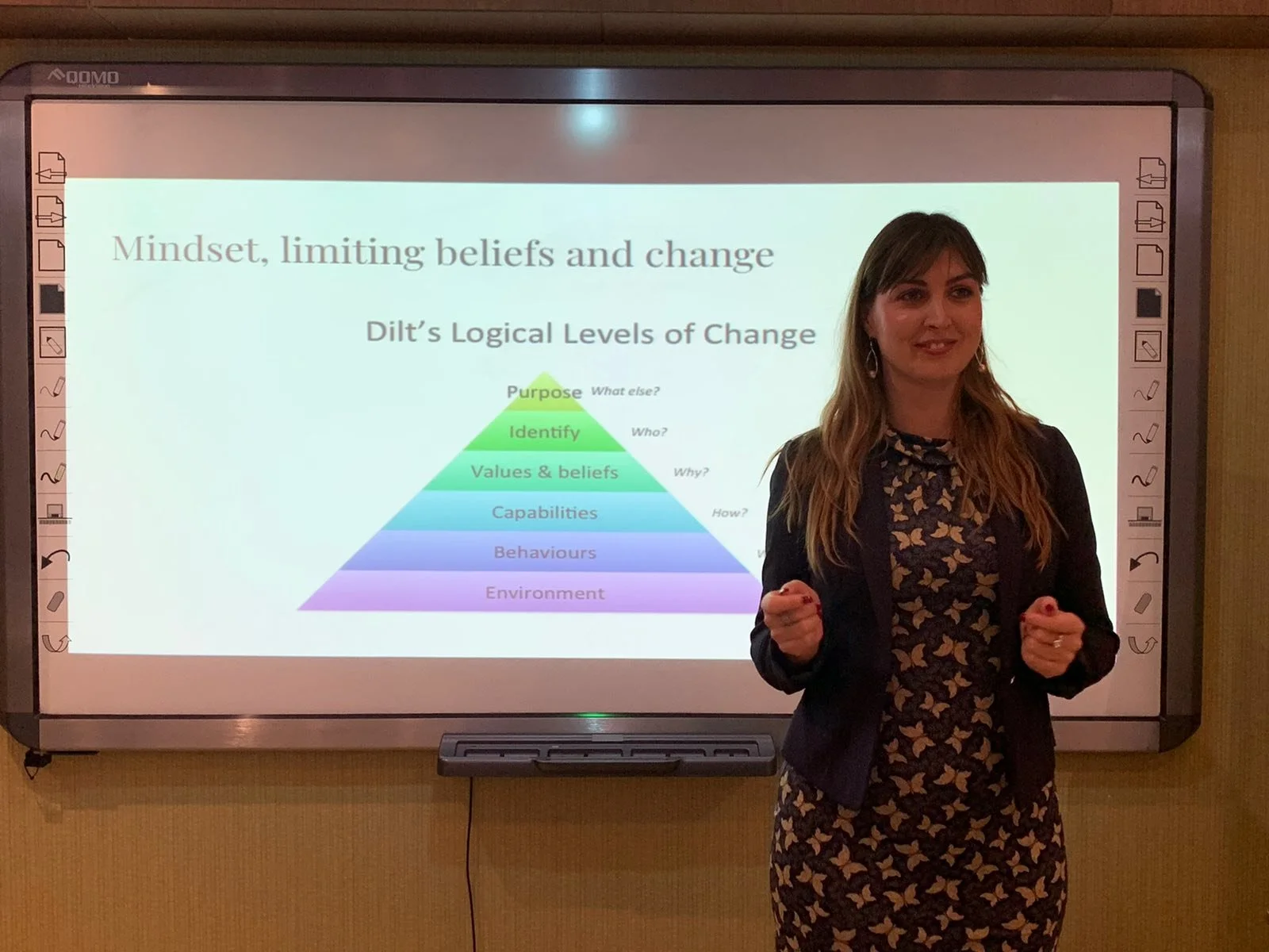 A woman standing in front of a presentation slide titled 'Mindset, limiting beliefs and change'. The slide features a colorful pyramid diagram labeled 'Dilt's Logical Levels of Change' with levels from bottom to top: Environment, Behaviours, Capabili