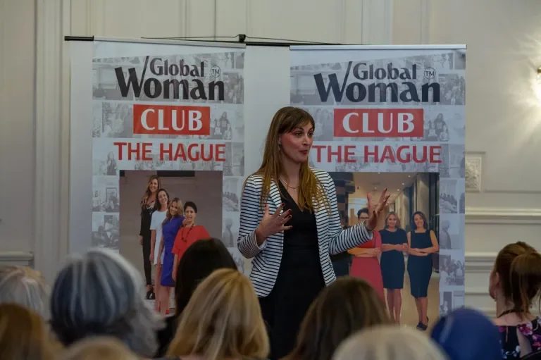 A woman giving a presentation at a speakers' event, standing in front of two banners that read 'Global Woman Club The Hague.' An audience is seated in front of her.