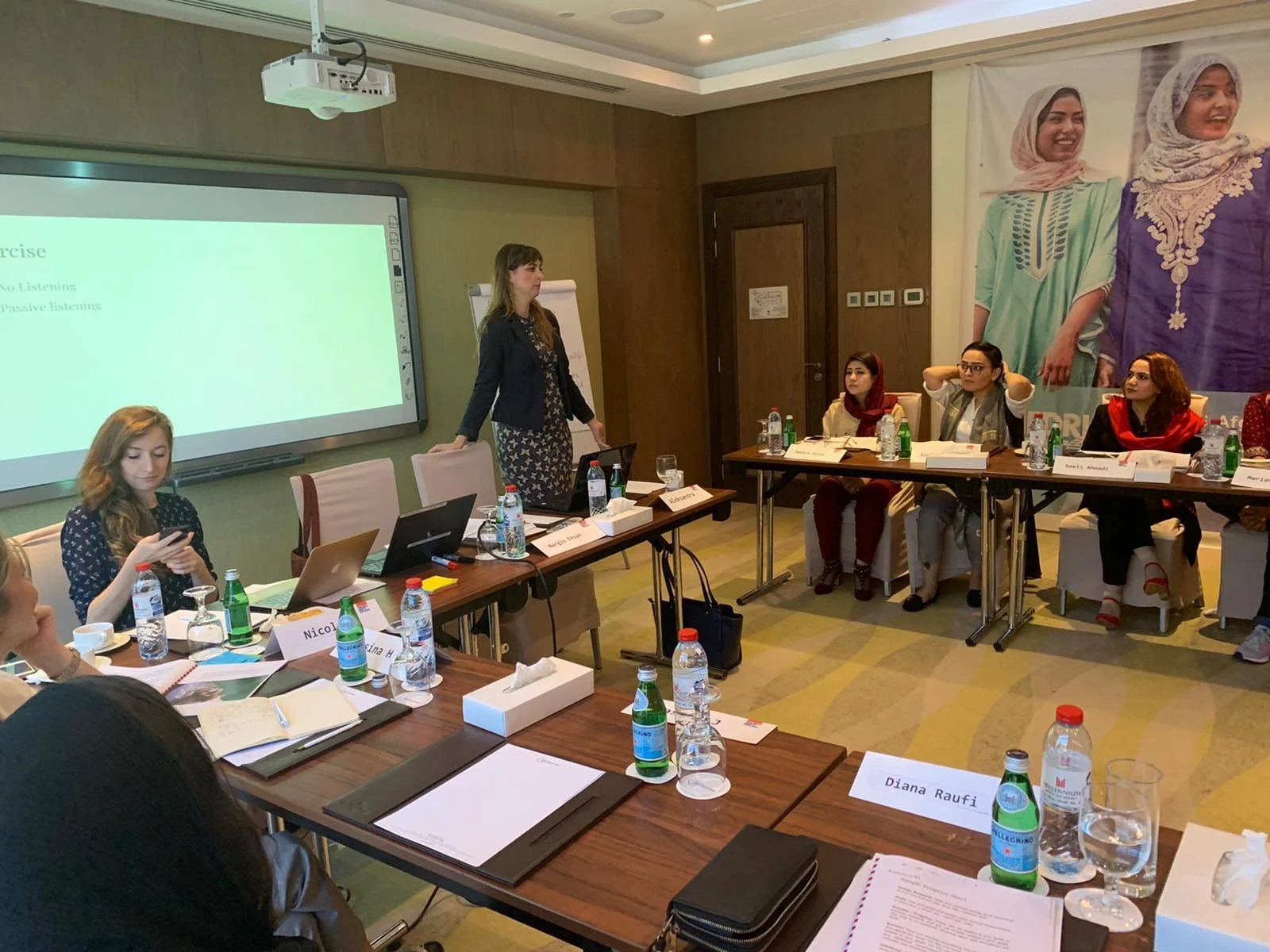 A group of women seated at a large conference table in a meeting room, with one woman standing and speaking. The table has notebooks, water bottles, glasses, and nameplates. A large poster of women in traditional attire and a digital screen with text