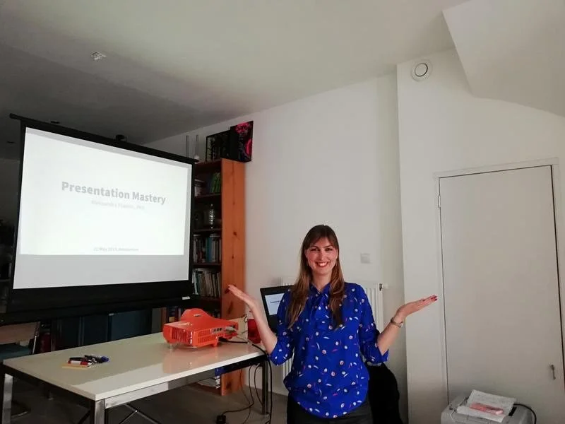 A woman standing in a room with a presentation slide on a screen behind her, smiling and gesturing with her hands, with a bookshelf and a closed white door in the background.