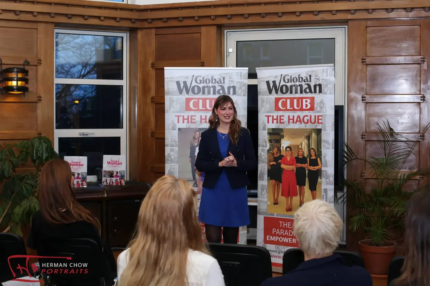 A woman speaking at a presentation or event, standing in front of two banners with the 'Global Woman Club The Hague' logo. Several seated audience members are visible, and the room has wooden walls and some potted plants.