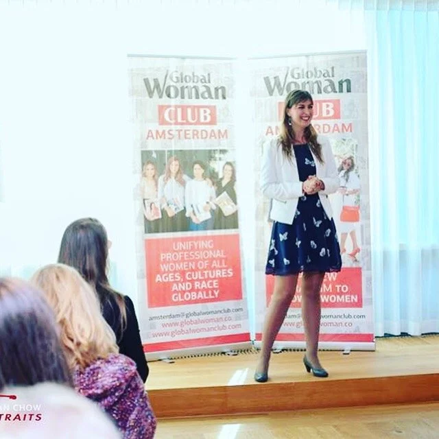 A woman in a white blazer and blue dress speaking at a Global Woman Club event in Amsterdam, standing in front of banners with the club's logo and text about unifying professional women worldwide.