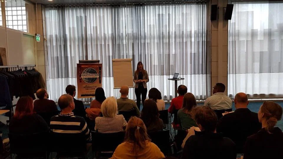 A woman standing in front of a group of seated people giving a presentation in a conference room with large windows and curtains.