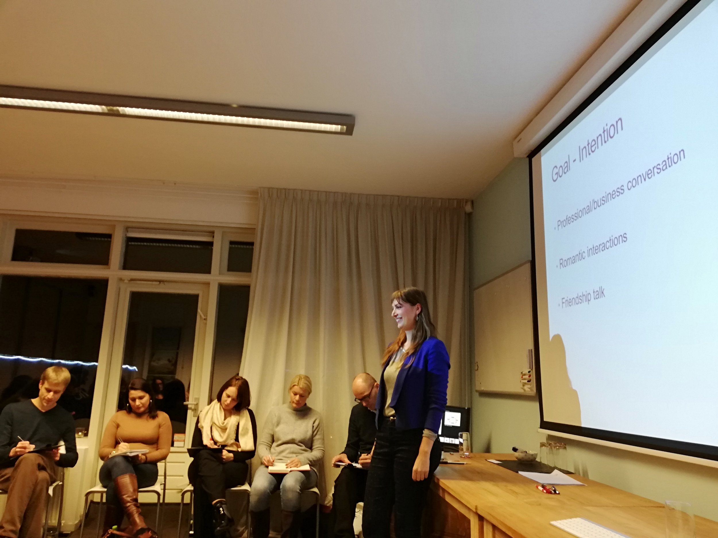 A woman in a blue blazer presenting in front of a projected slide titled 'Goal - Intention' at a group meeting with people seated and taking notes in a well-lit room with large windows and beige curtains.