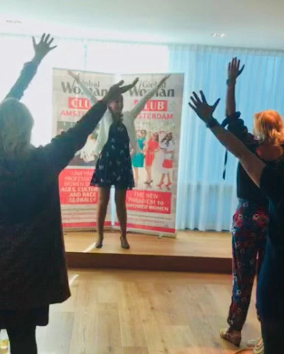 Women participating in a dance class with arms raised, inside a room with a banner in the background.