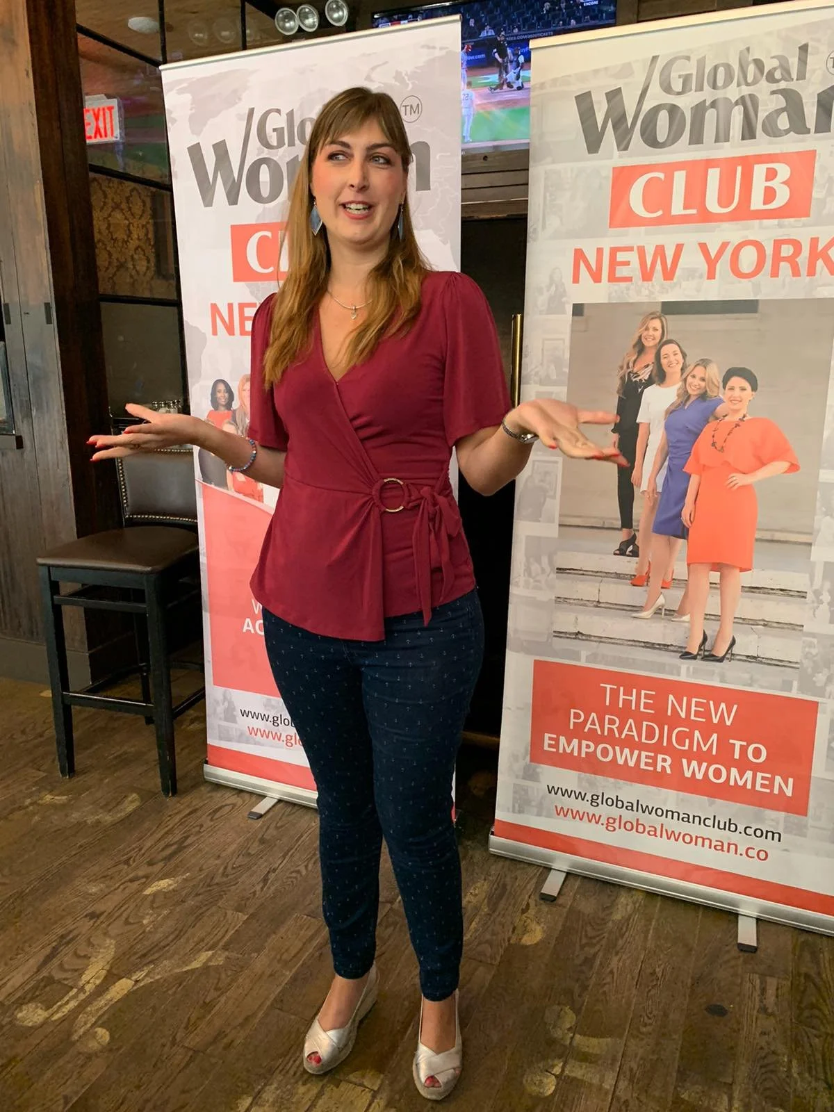 A woman with long light brown hair, wearing a red wrap top and dark blue polka dot pants, standing in front of two posters for Global Women Club New York, with her hands raised and a smile.