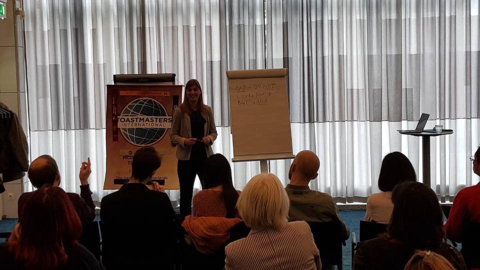 Woman giving a presentation at Toastmasters event, standing in front of a flip chart and Banner, audience seated facing her, with a laptop and glass of water on a table to her right.