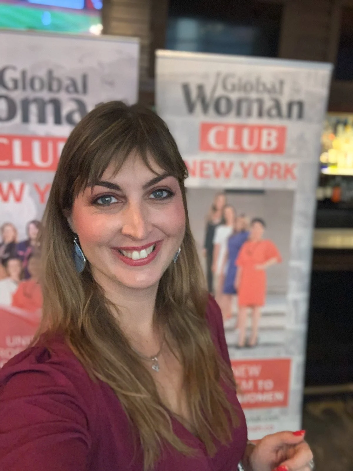 A woman taking a selfie in front of a banner that reads 'Global Woman Club New York' with a group of women pictured below the text.