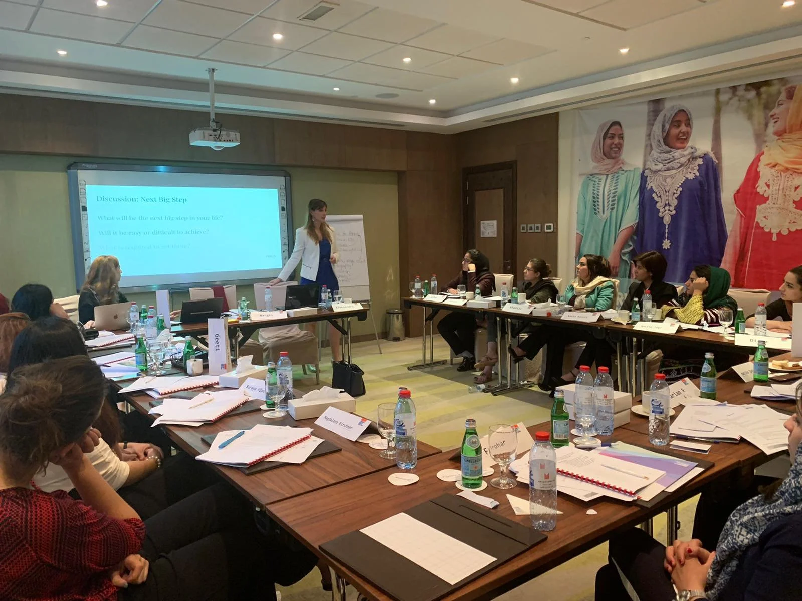 A woman is standing and presenting during a business meeting or workshop, with attendees seated at a U-shaped table, various documents, water bottles, and nameplates visible, in a conference room with large wall art of women in traditional attire.