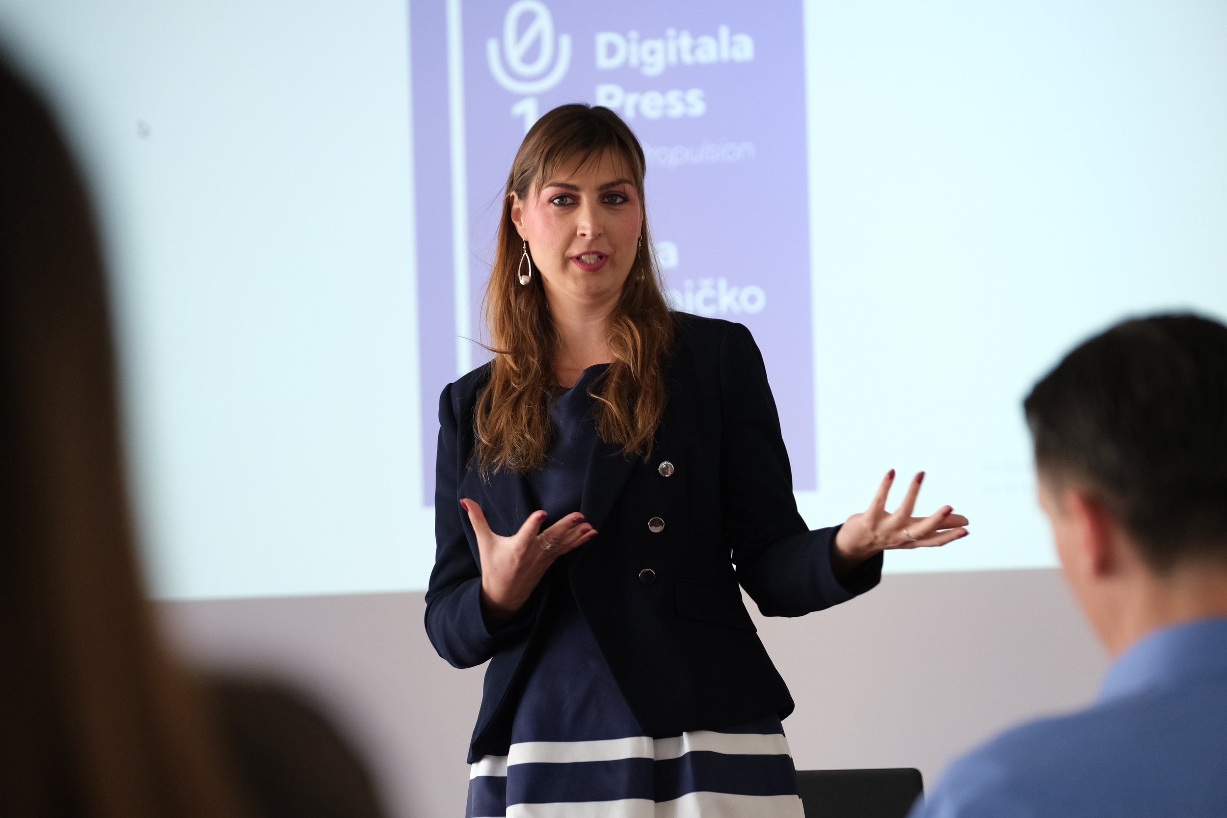 A woman with brown hair and earrings speaking or presenting in front of a white screen with a purple graphic, during a presentation or lecture, with audience members visible in the foreground.