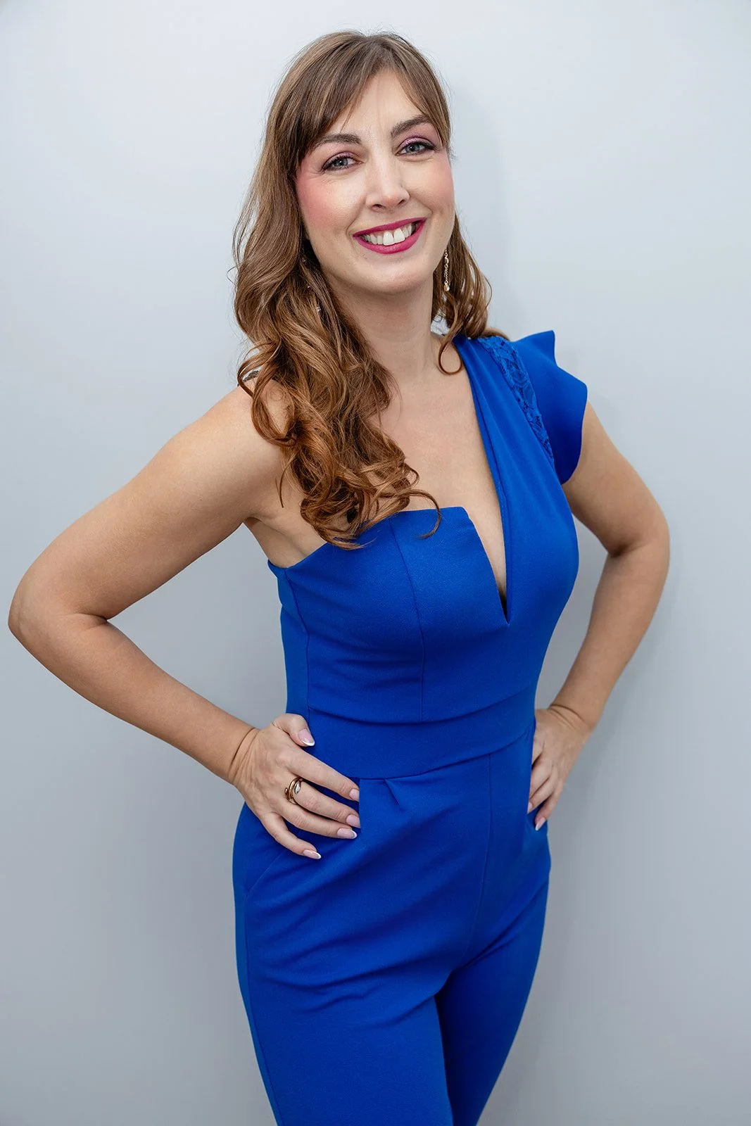 A woman with long wavy brown hair wearing a bright blue jumpsuit, standing with her hands on her hips and smiling.