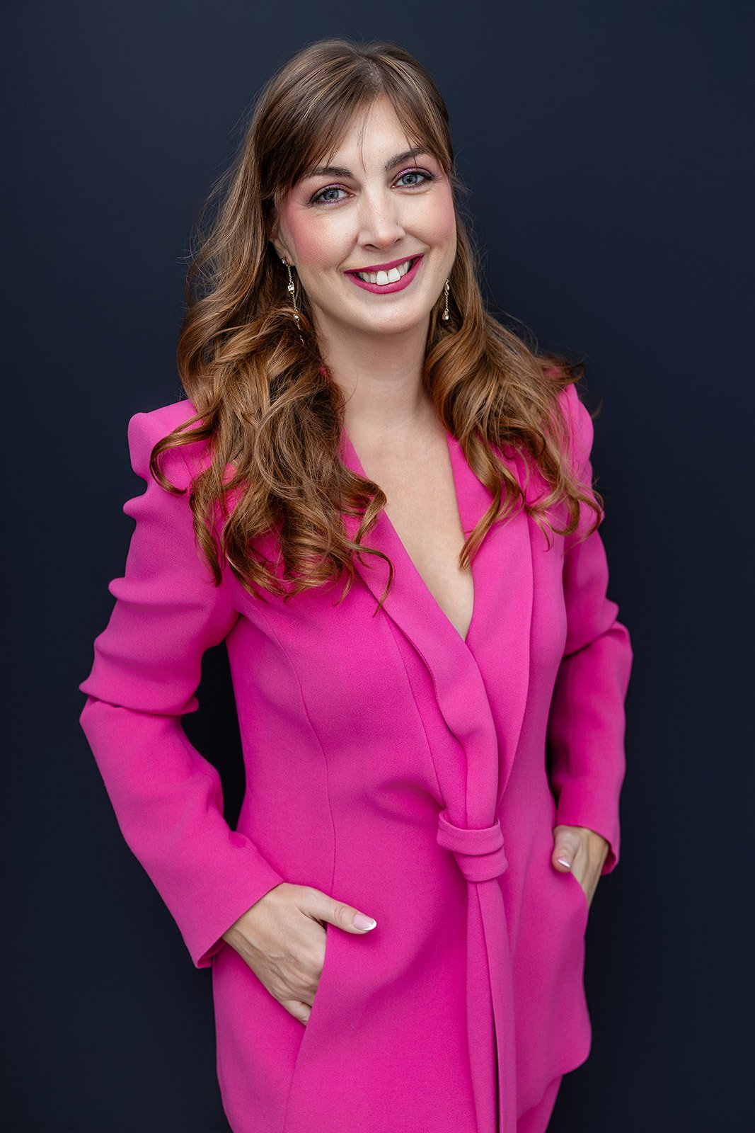 A woman with long wavy brown hair, wearing a bright pink blazer, smiling and standing against a dark background.