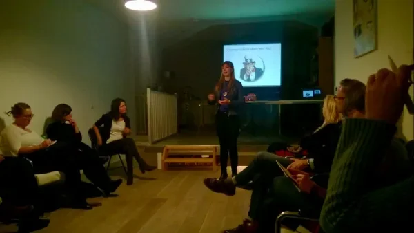 A woman is giving a presentation to a group of people in a dimly lit room, with a screen displaying a slide in the background.