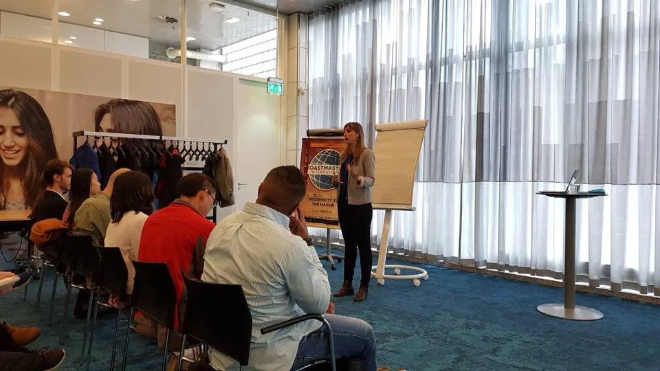 A woman is giving a presentation to a group of people seated in a modern conference room with large windows and sheer curtains.