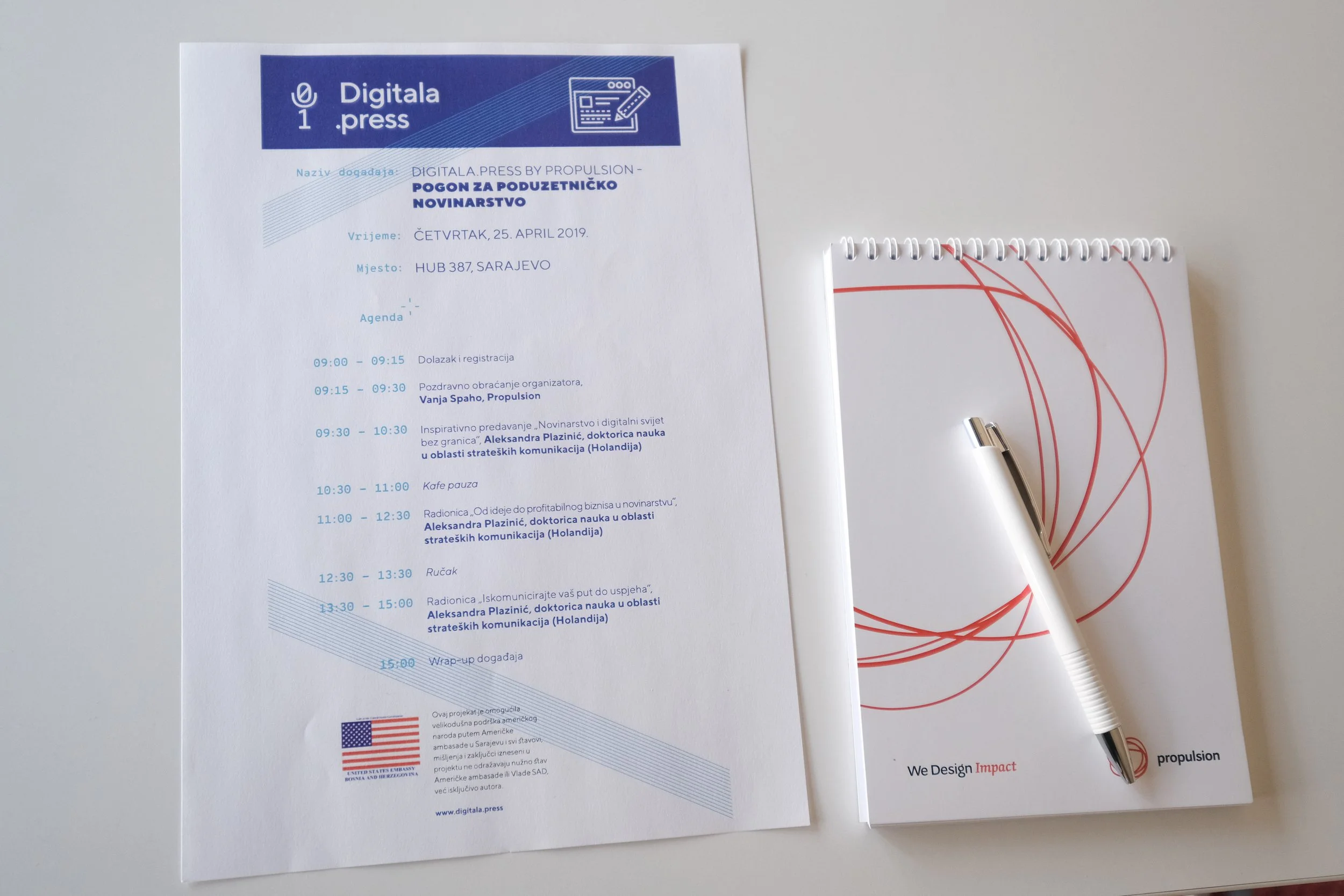 Event schedule printed on a white sheet of paper with blue accents, placed on a white surface. To the right, a pen with red wires attached to it and a small white device are visible.