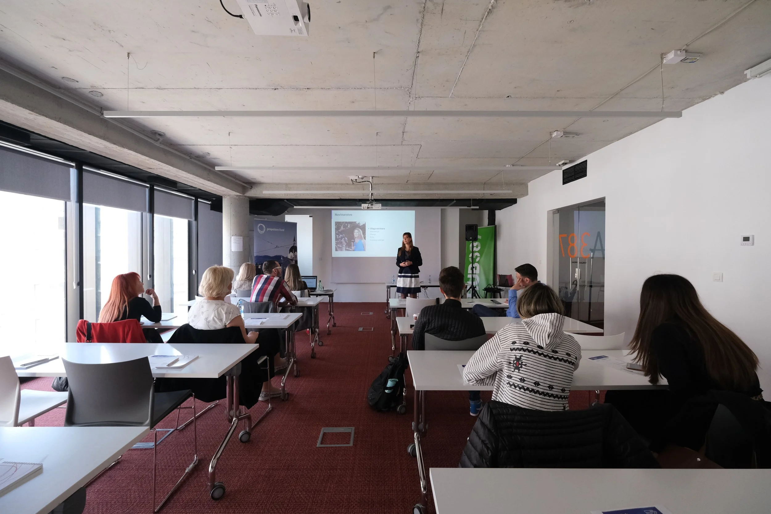 A woman giving a presentation in a conference room with audience members seated at tables, a projection screen displaying a slide, and large windows.