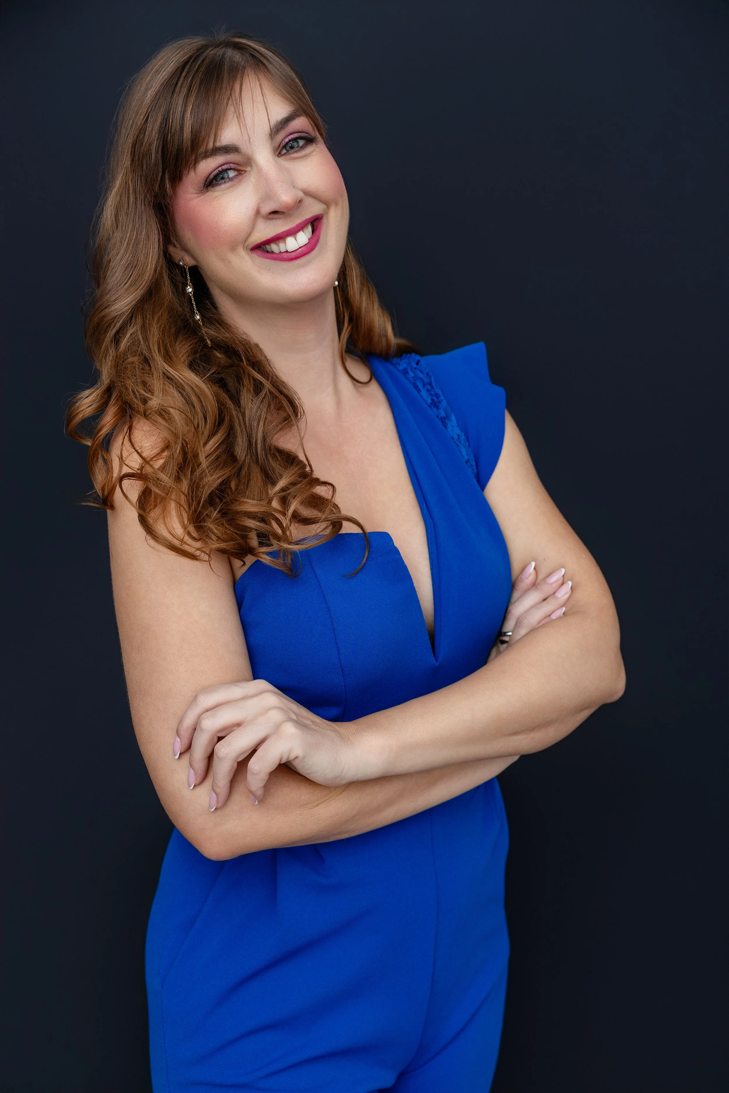 A woman with long wavy red hair, wearing a blue dress, standing against a dark background, smiling with crossed arms.
