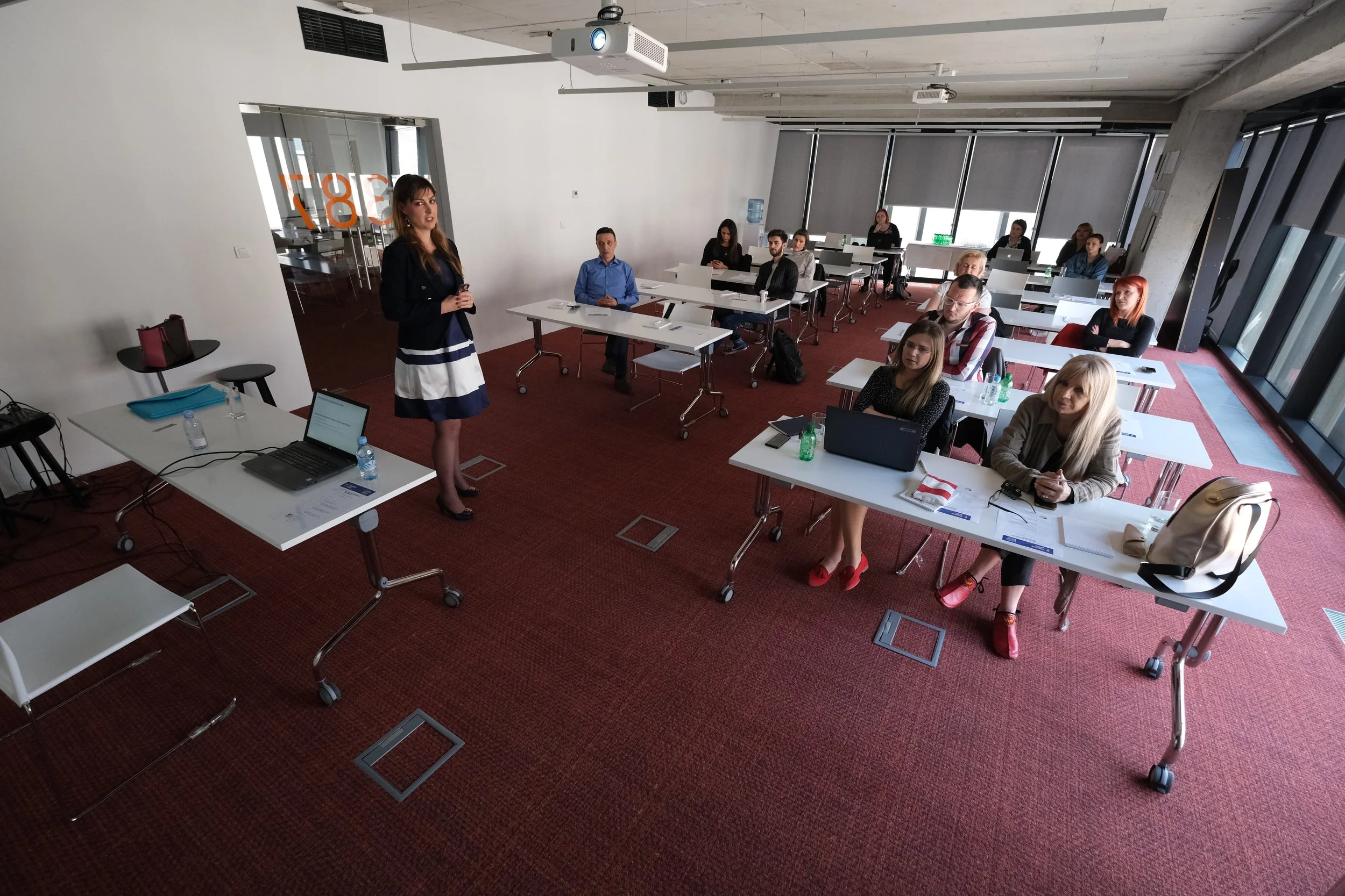 A woman in a black blazer and striped dress giving a presentation in a modern conference room. Attendees are seated at white tables, some with laptops, water bottles, or notebooks. The room has floor-to-ceiling windows with blinds, a projector mounte