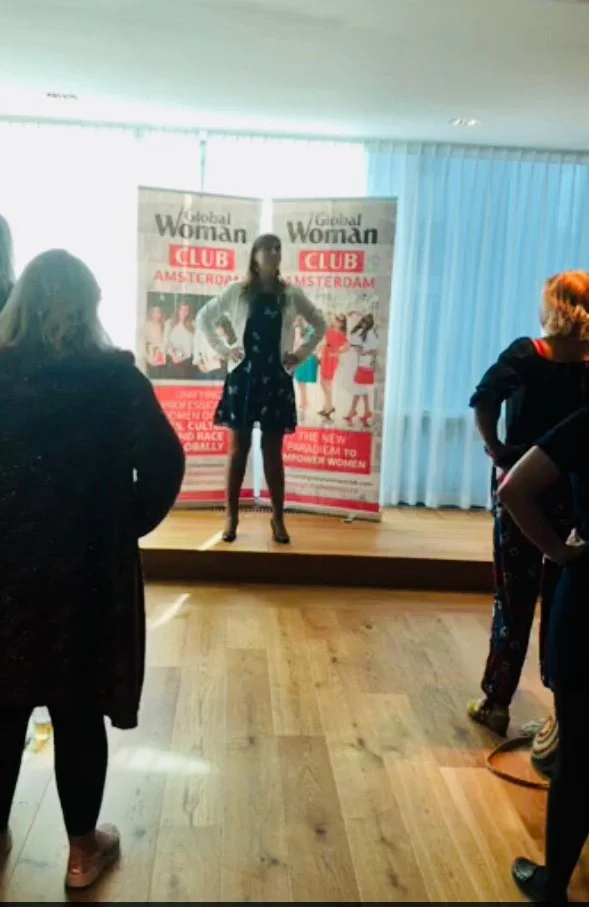 A woman standing on a small stage with two banners behind her reading 'Global Woman CLUB Amsterdam'. She is wearing a black dress with a floral pattern and a white jacket. Several women are standing in front of the stage.