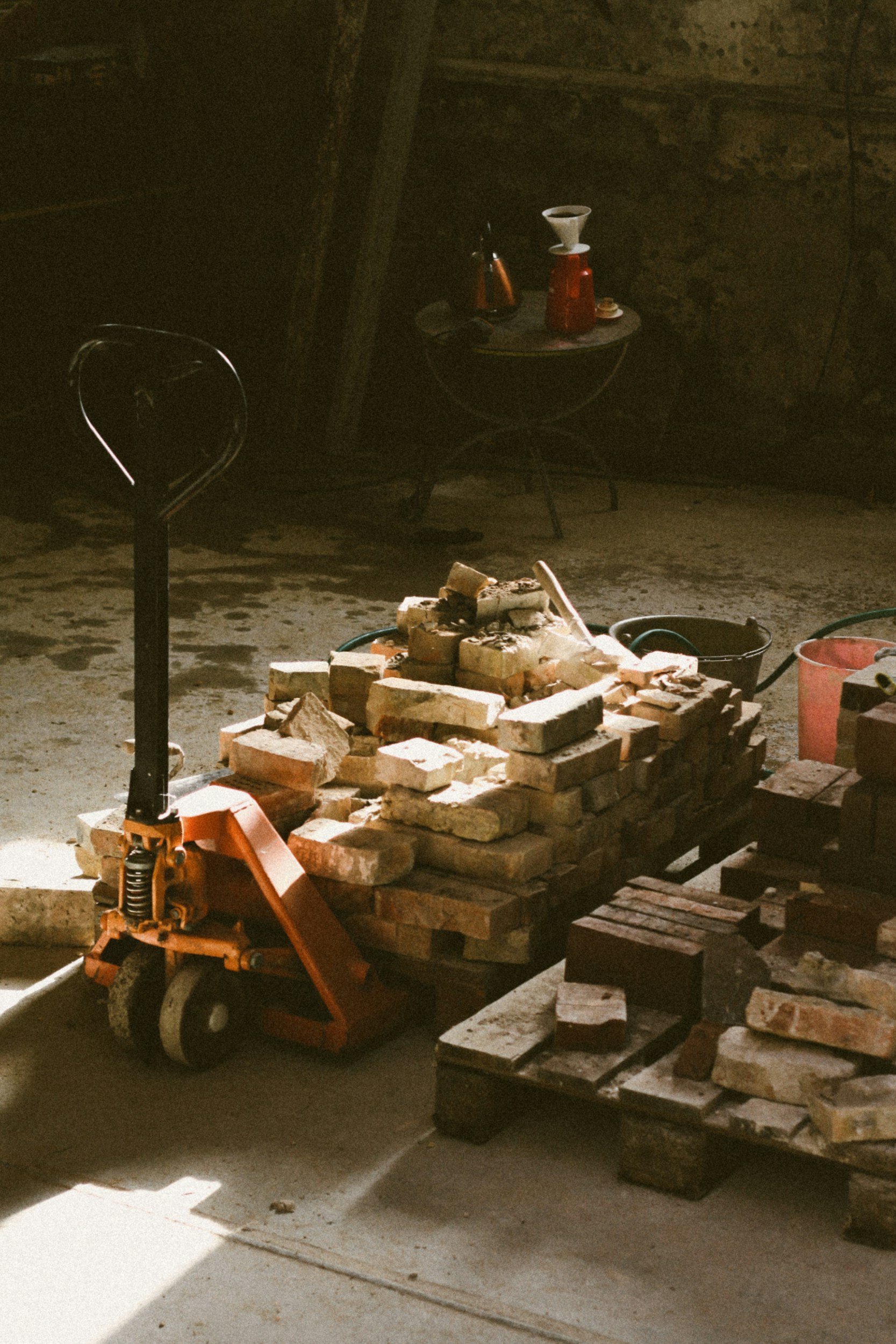 A brick pile on a hand truck with a small table holding bottles and a coffee dripper on a wall in a dimly lit room, with construction materials and equipment around.
