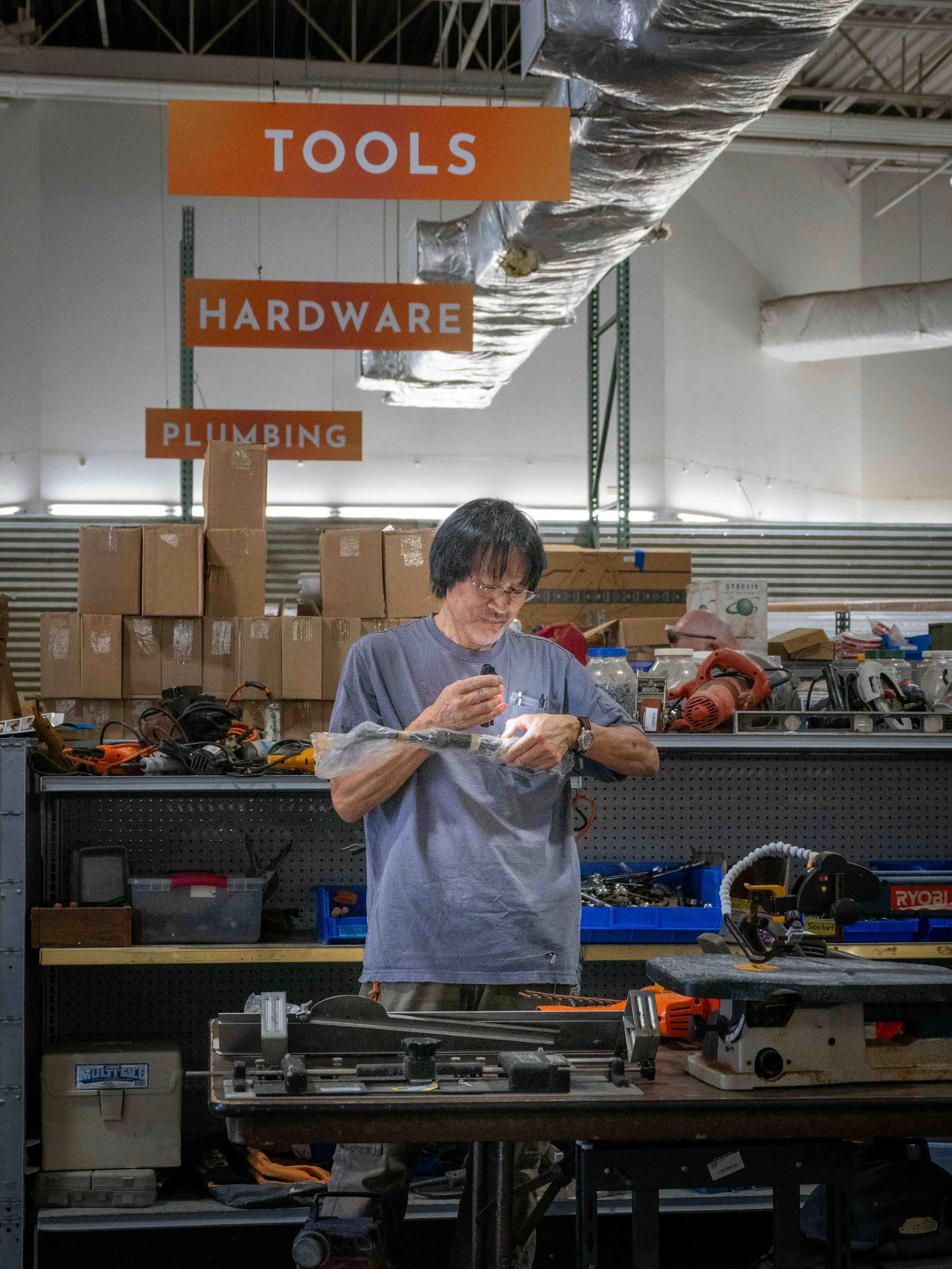A man working in a hardware store or workshop, surrounded by tools and equipment, with signs indicating sections for tools, hardware, and plumbing.