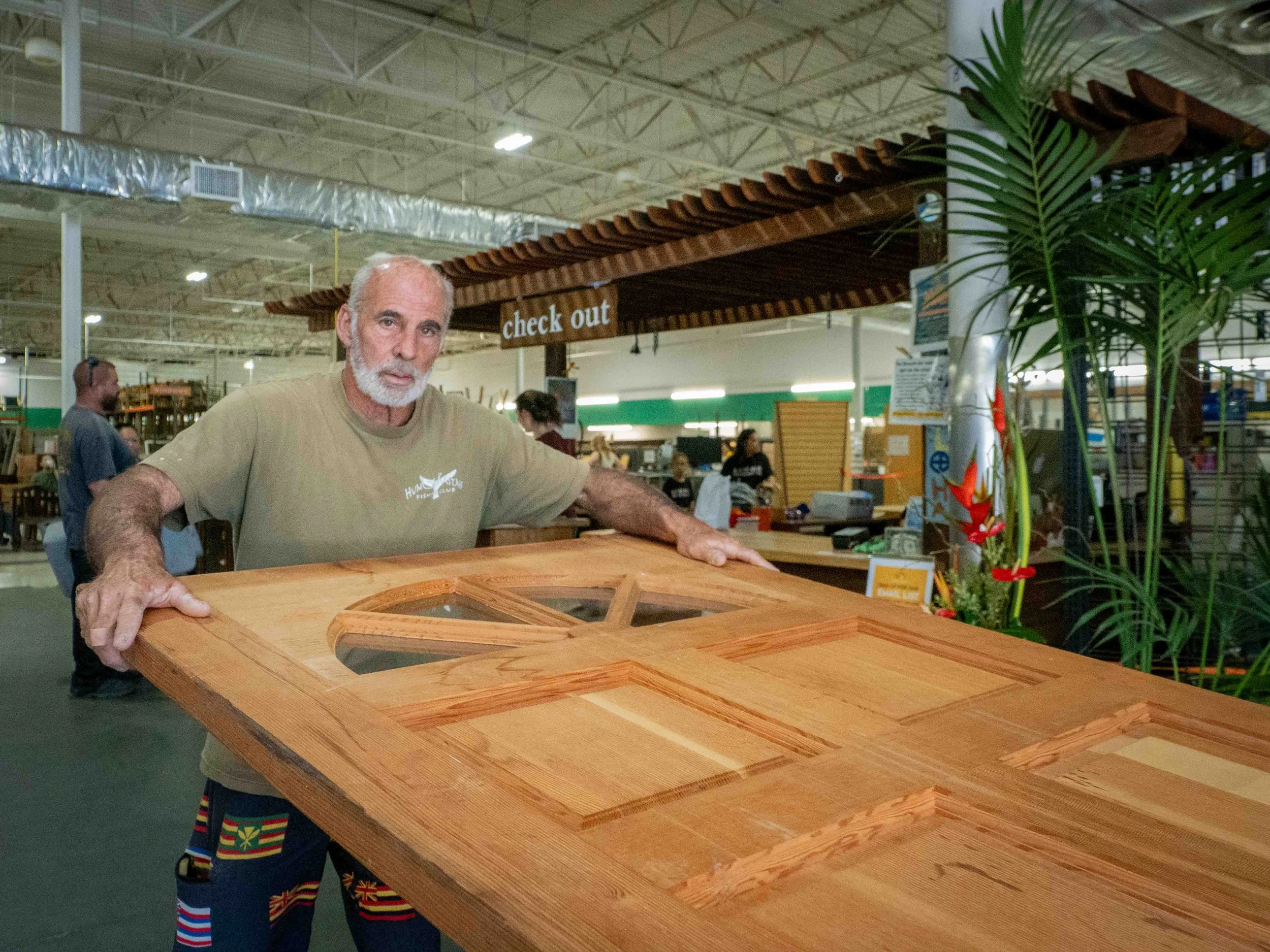 An elderly man with gray hair and a beard holding a large wooden table with glass inserts inside a furniture store or warehouse.